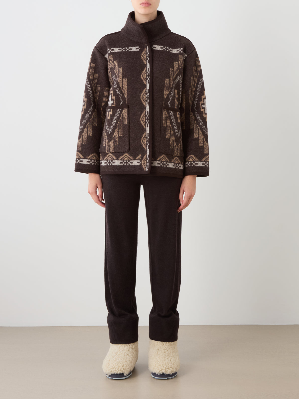 A person wearing a Pioneer Jacket women in an oversized boiled wool with a geometric pattern, matching brown trousers, and cream-colored fleece slippers.
