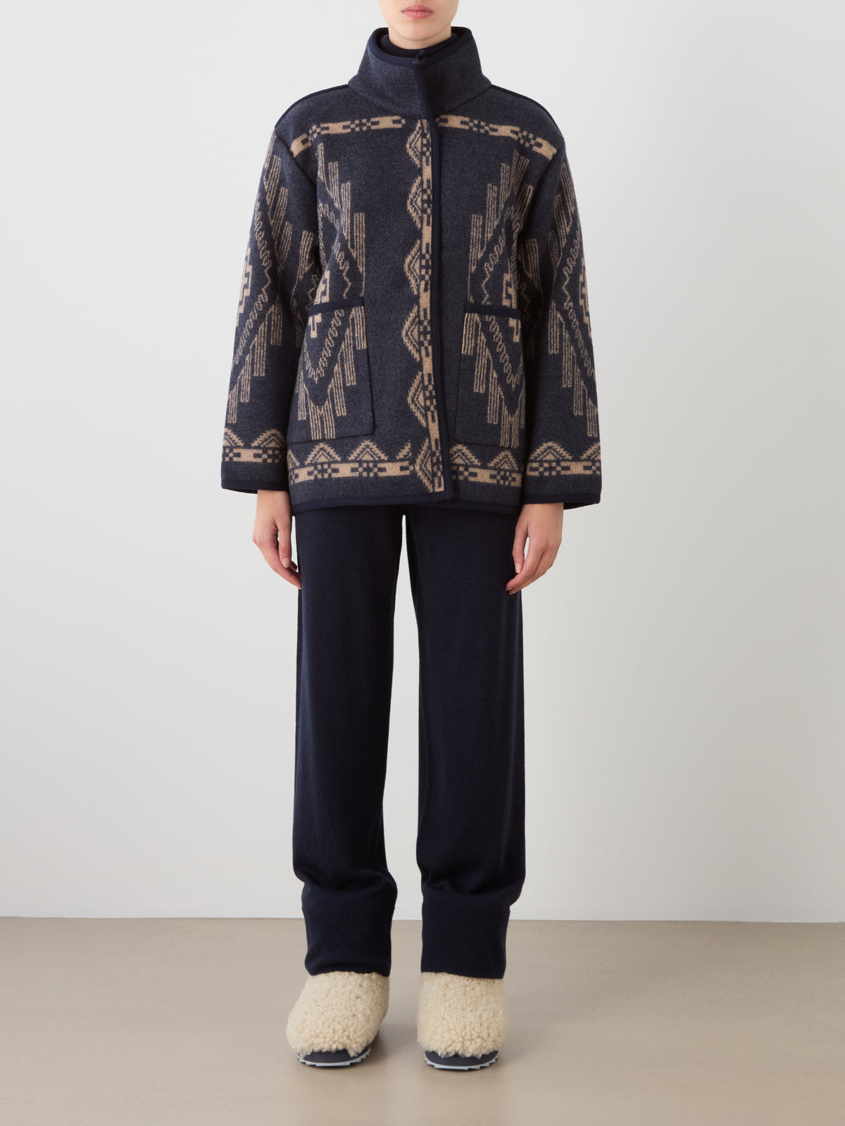 An oversized navy blue Pioneer Jacket women in a wool-blend with a tan geometric pattern, worn over matching navy trousers and cream-colored fleece-lined shoes.