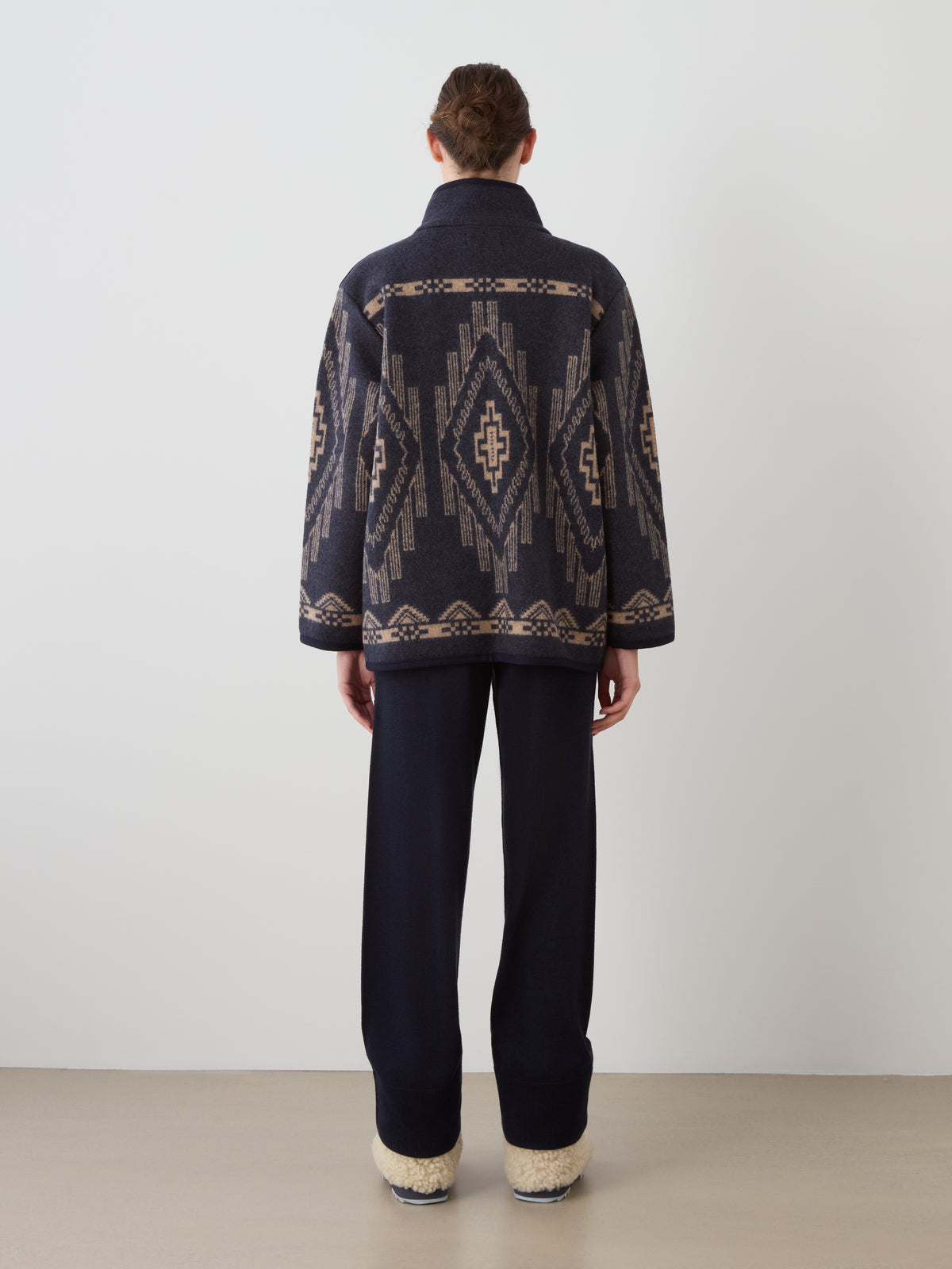 A person viewed from behind wearing a Pioneer Jacket women with a tan geometric pattern, dark pants, and light-colored fuzzy slip-on shoes.