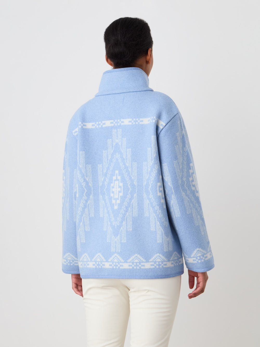 A person with dark hair tied back models the We Norwegians Pioneer Jacket women - Pre Spring 26 in light blue with white geometric patterns and white pants, standing with their back to the camera against a plain light background.