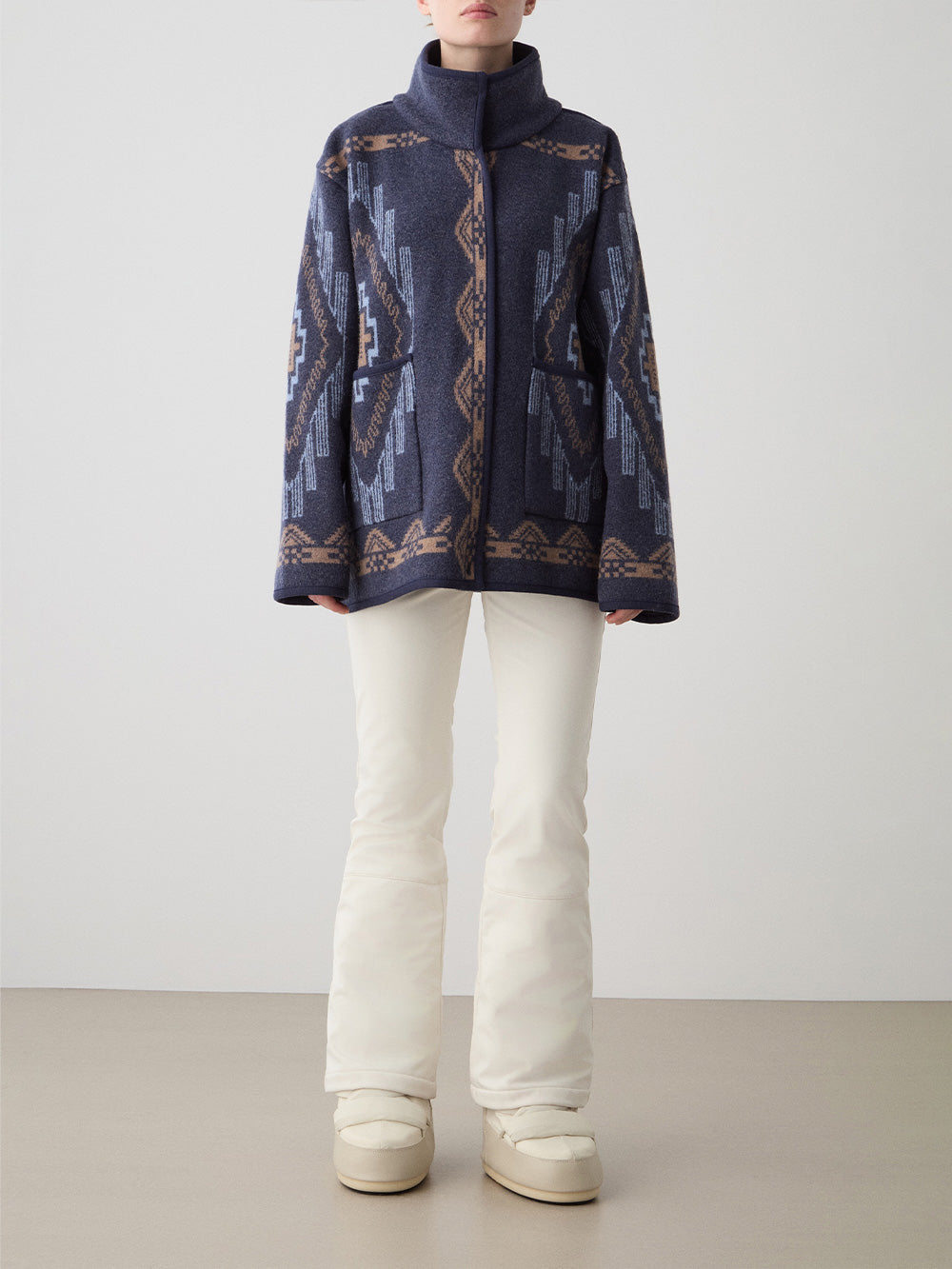 A person wears the We Norwegians Kautokeino Jacket for women—a blue and brown jacquard-patterned boiled wool jacket with a high collar—paired with white flared pants and boots, standing against a plain, light background.