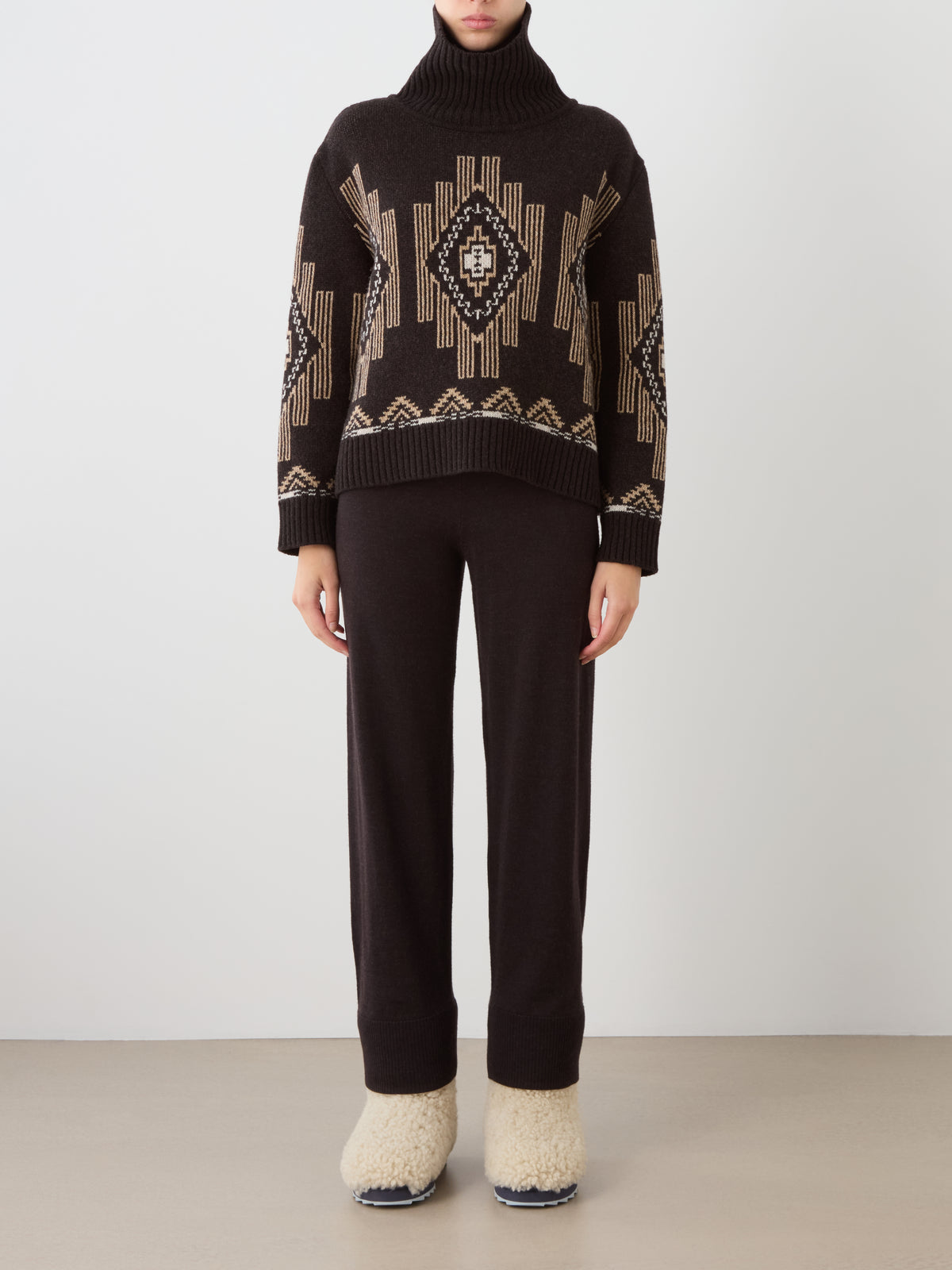 A person wearing a brown We Norwegians Pioneer Sweater women featuring a geometric pattern in tan and white, paired with matching lounge pants and beige shearling-lined slip-on shoes.