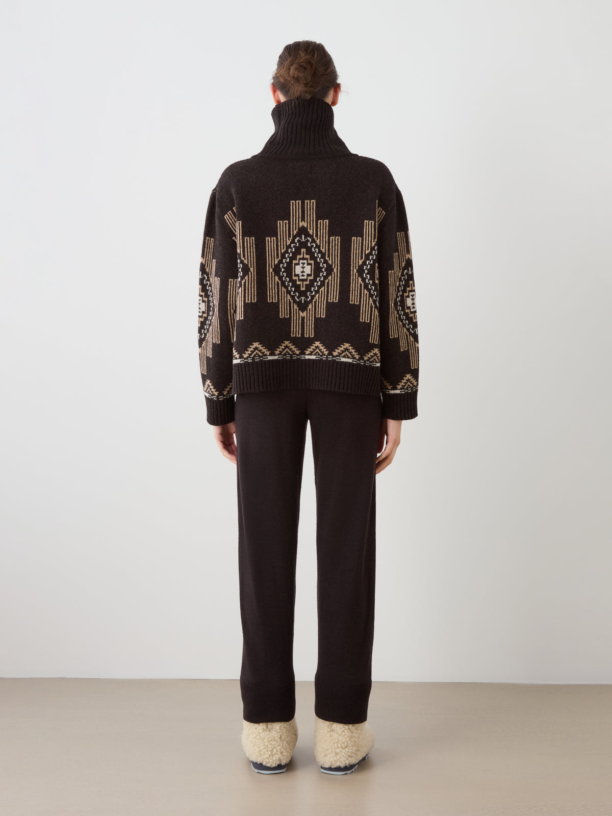 Back view of a person wearing a dark brown wool blend Pioneer Sweater women with a geometric beige and cream pattern, paired with matching dark brown trousers and beige slippers.