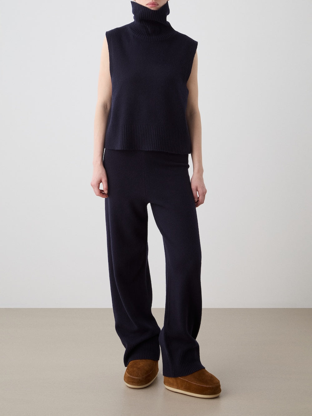 A person stands against a plain background wearing We Norwegians Geilo Jogger women set in navy, featuring a relaxed fit sleeveless turtleneck sweater and wide-leg pants, styled with brown suede slippers. Their arms are at their sides, face partly covered by the turtleneck.