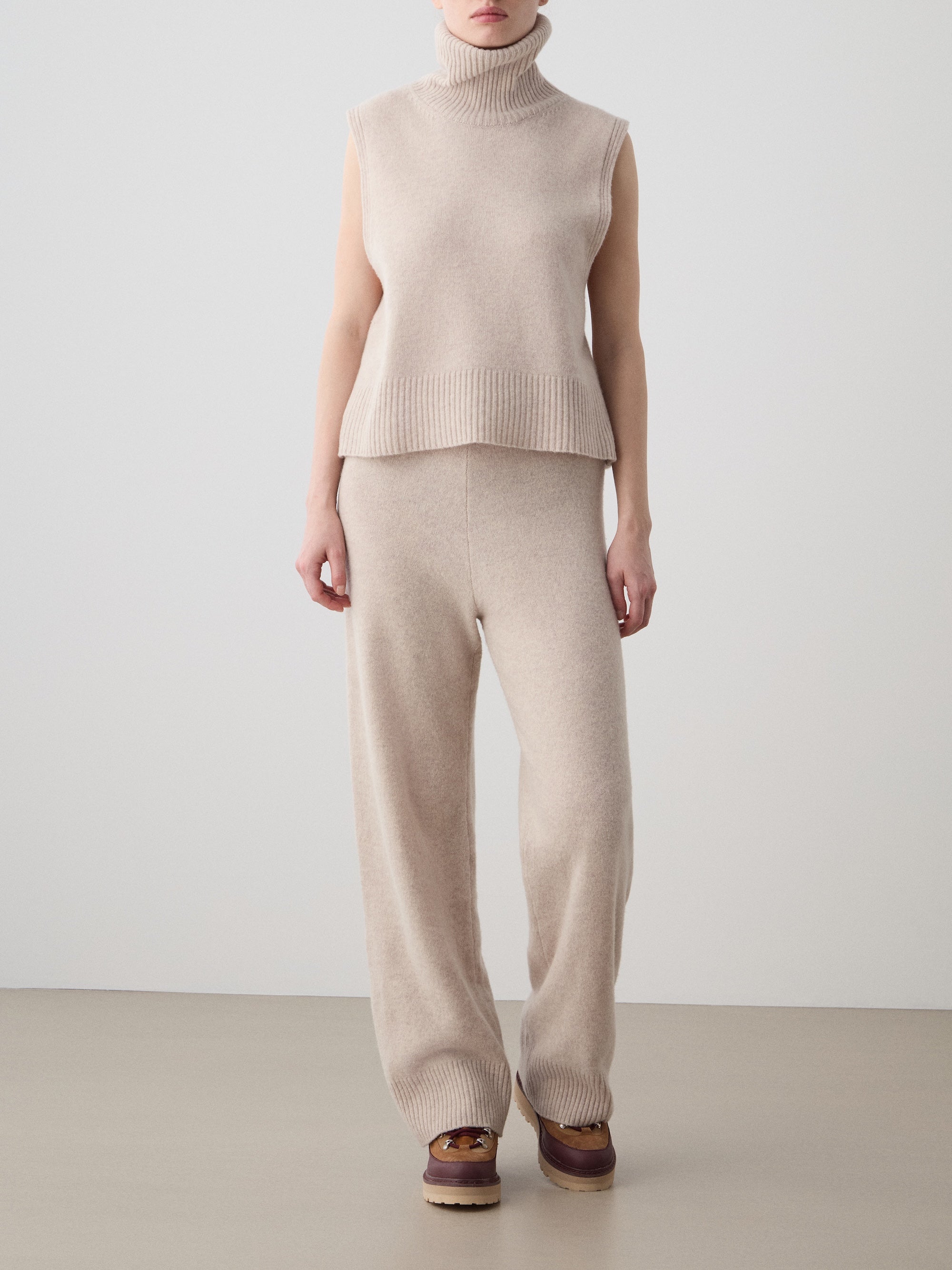 A woman wearing We Norwegians' Geilo Jogger stands against a plain light background, styled with a beige sleeveless turtleneck sweater and brown slip-on shoes.