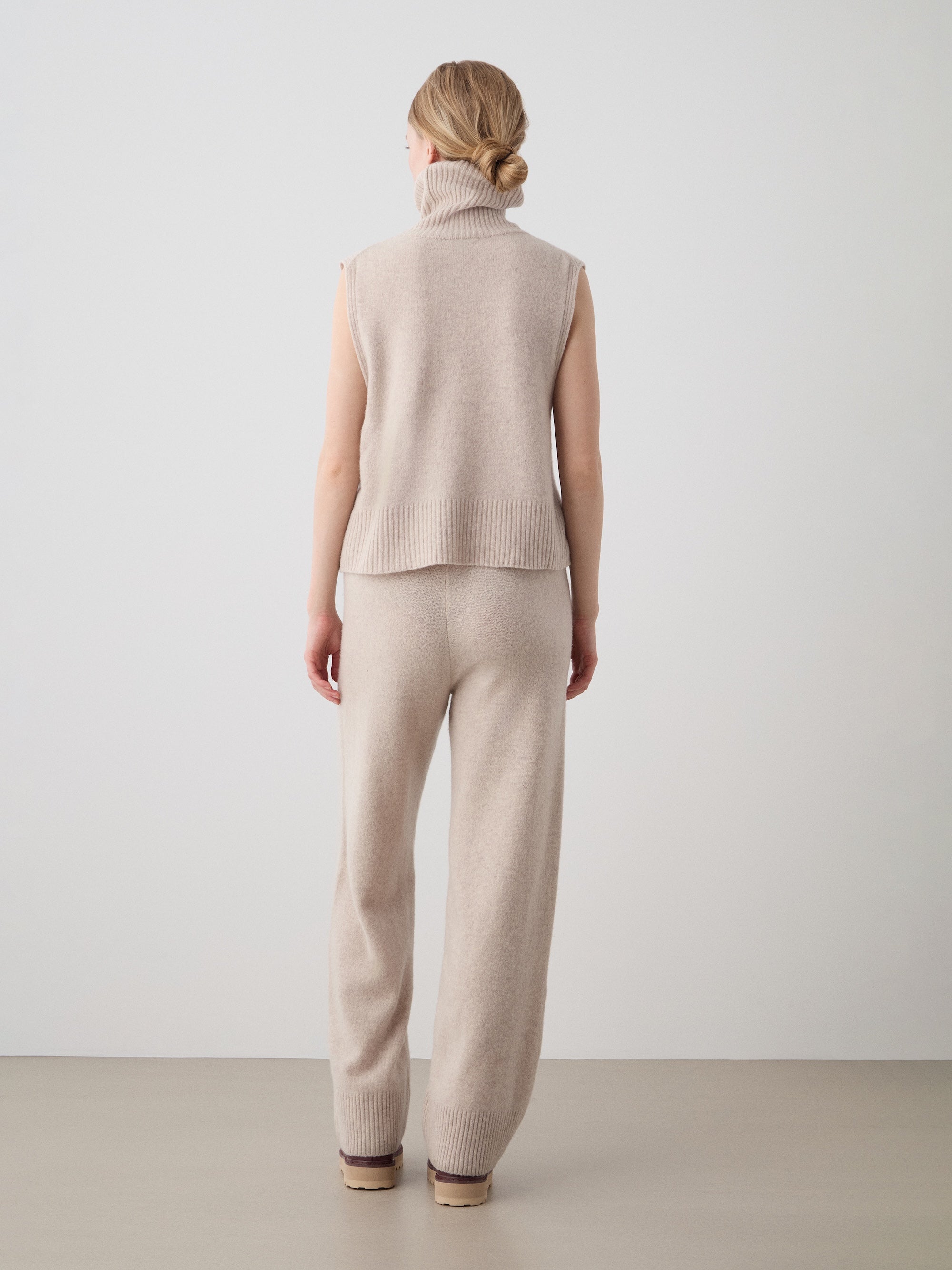 A woman with blonde hair in a low bun, wearing a sleeveless beige turtleneck and We Norwegians Geilo Jogger women’s knit pants, stands indoors facing an off-white wall with her back to the camera.