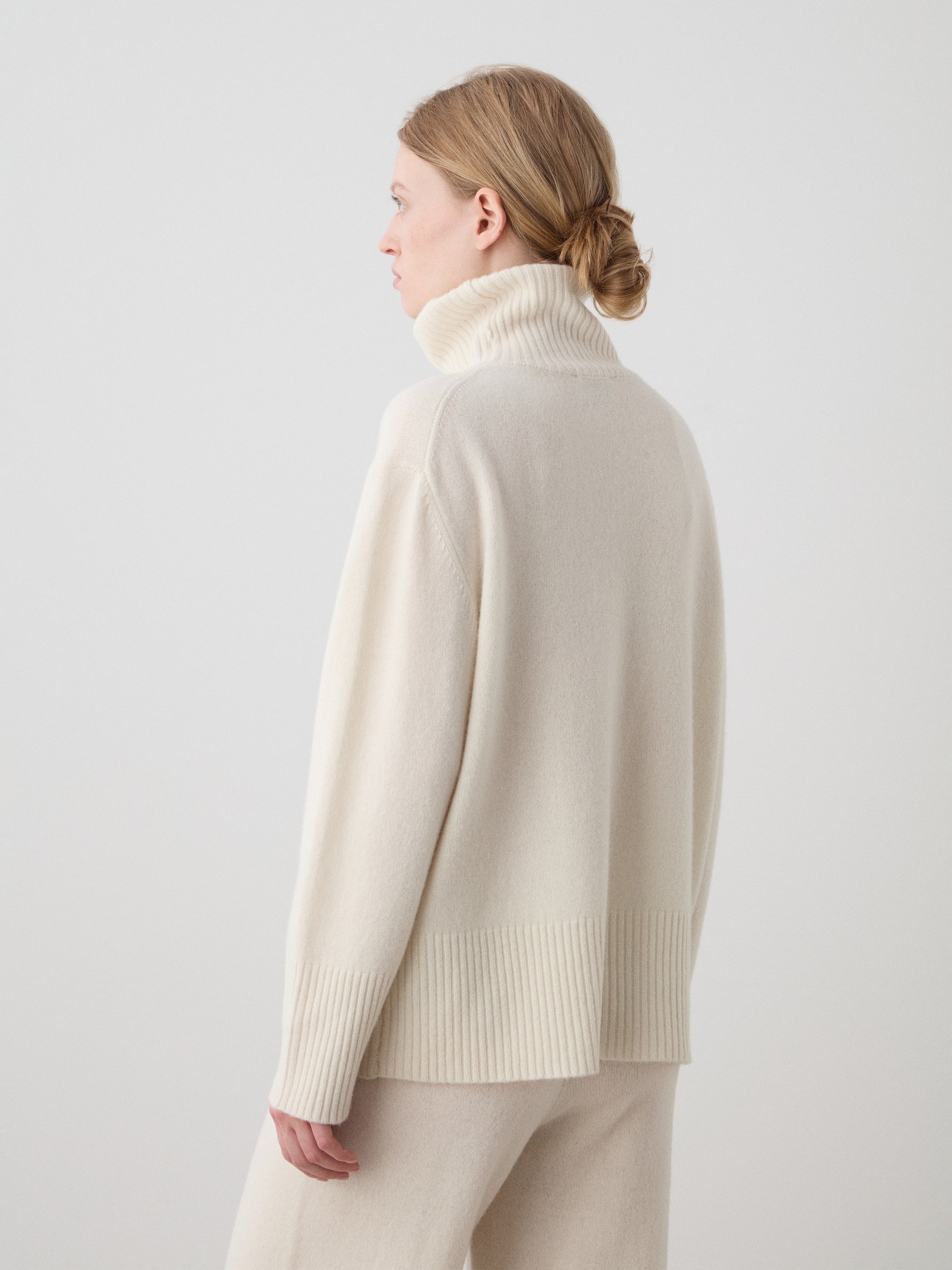 A woman with light brown hair in a low bun wears the We Norwegians Geilo BoyFriend Sweater for women, made from Extrafine Merino Wool, standing against a plain light background and facing away from the camera.
