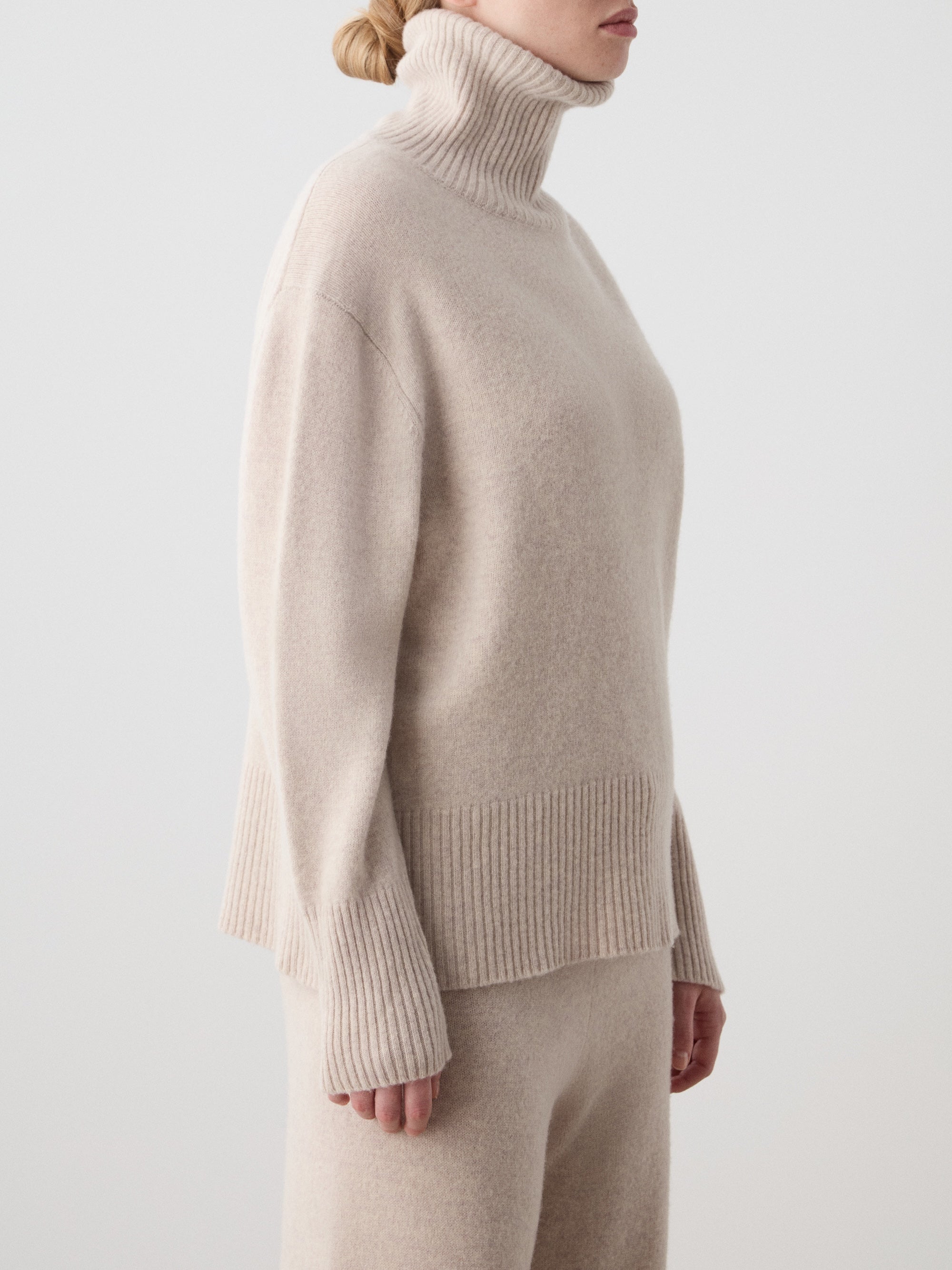 A person wears the We Norwegians Geilo BoyFriend Sweater for women in beige extrafine Merino wool with ribbed cuffs and hem, paired with matching pants, against a plain light background. Their face is partially out of frame.