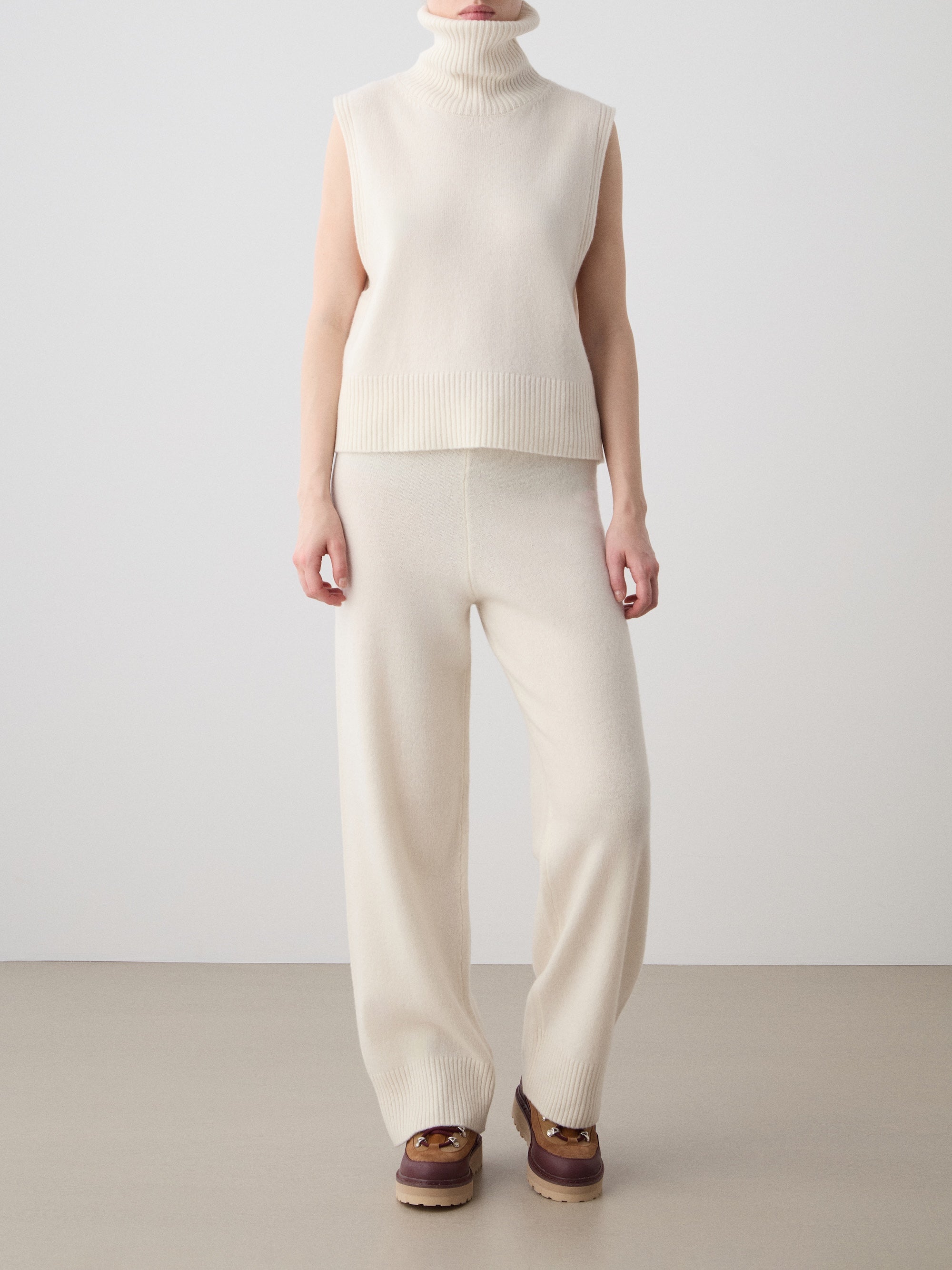 A person wears the We Norwegians Geilo Vest for women in cream, made from extrafine merino wool, paired with matching wide-leg knit pants and brown loafers, standing against a plain background.