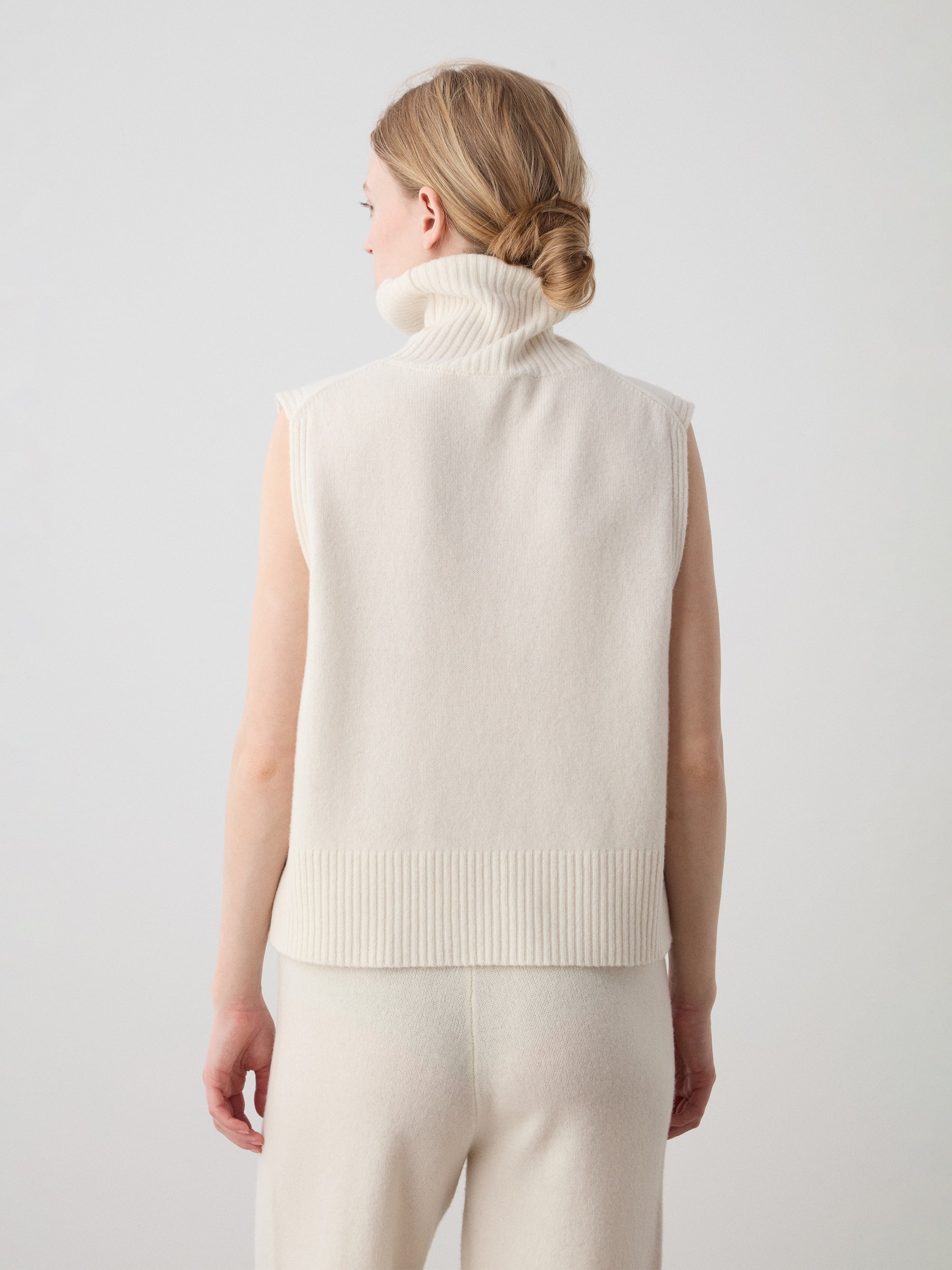 A woman with light hair, in We Norwegians’ Geilo Vest women—sleeveless, high-neck, cream extrafine merino wool—and matching pants, stands facing away against a plain light background.