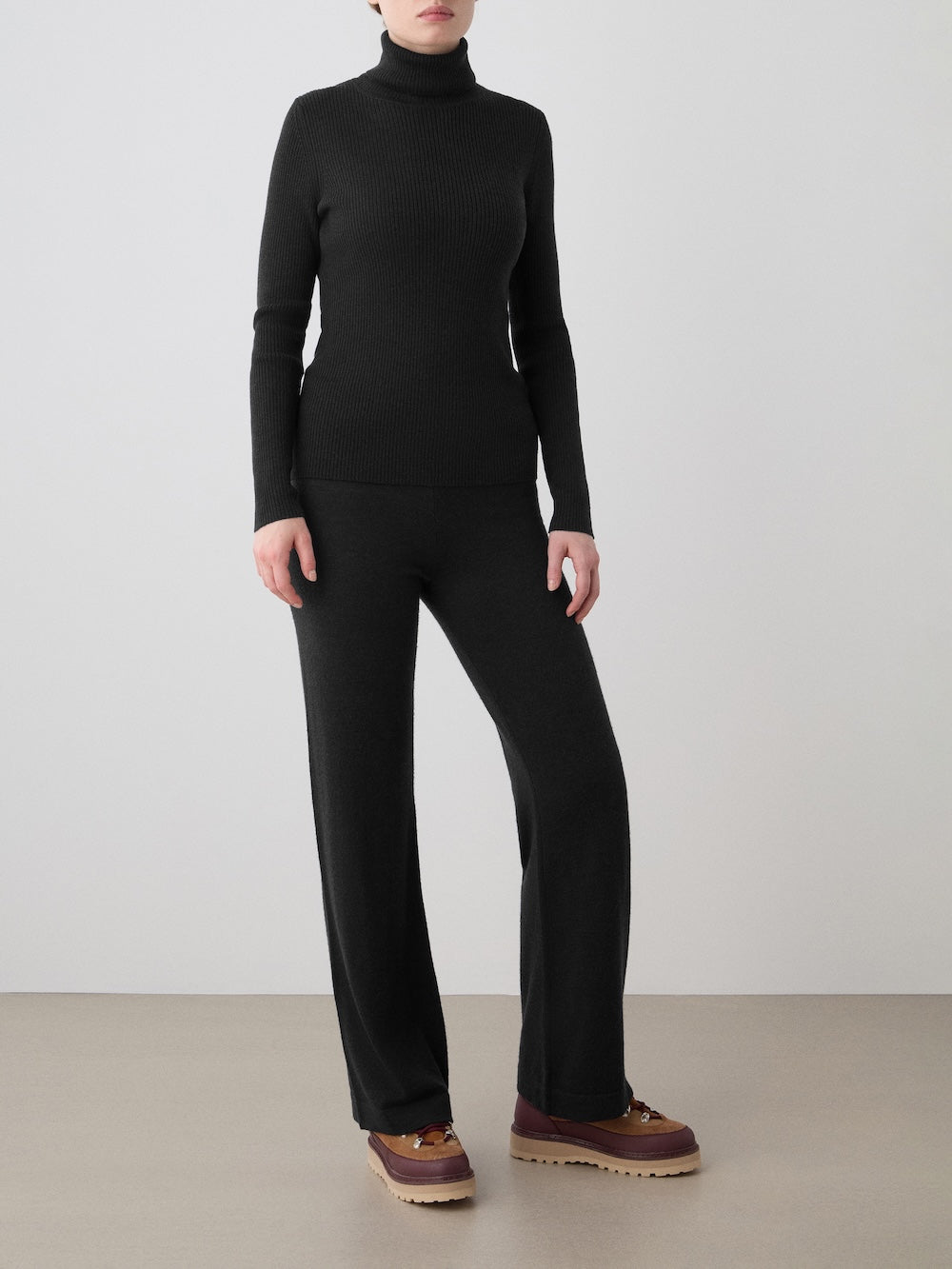 A person stands against a plain backdrop in the We Norwegians Geilo TurtleNeck women sweater, black wide-leg pants, and brown platform shoes. Their face is not visible.