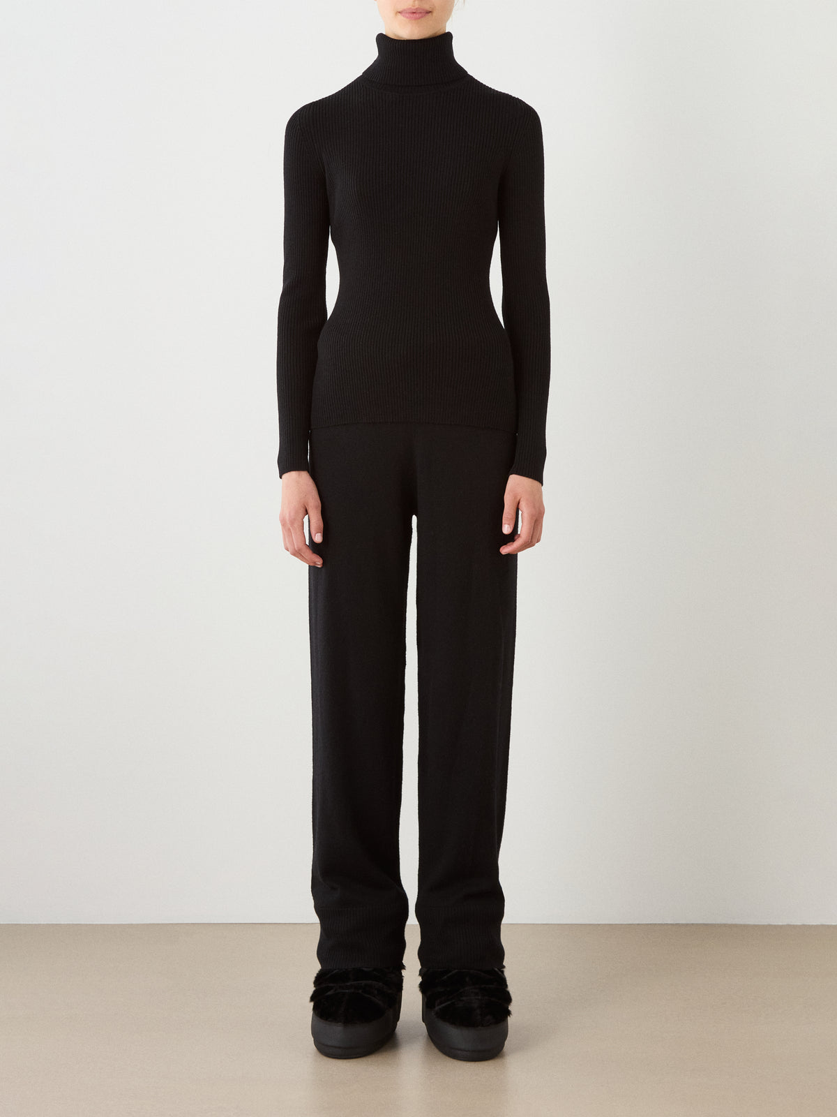 A person stands against a plain background wearing the We Norwegians Geilo TurtleNeck for women, paired with loose black pants and black shoes. Their arms are at their sides, and their face is not visible.