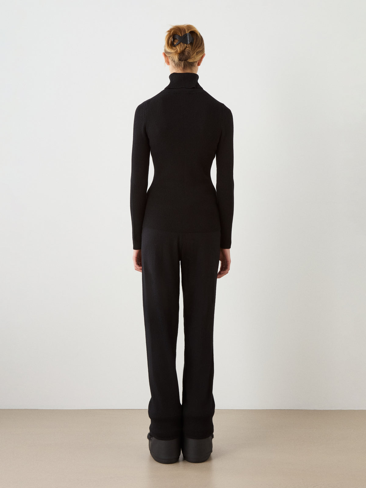 A person with blonde hair styled in an updo, dressed in the We Norwegians Geilo TurtleNeck women’s sweater, black pants, and black platform shoes stands against a plain white wall, facing away from the camera.