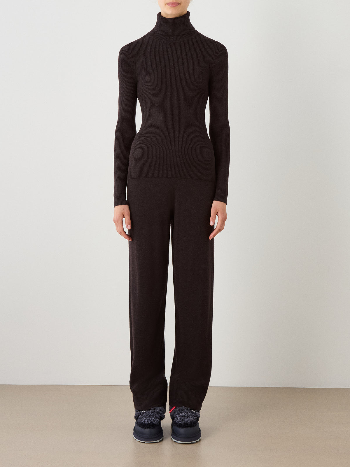 A person stands against a plain background wearing the We Norwegians Geilo TurtleNeck for women in black, paired with matching black wide-leg pants and dark shoes. Their hands are by their sides, and their face is not visible.