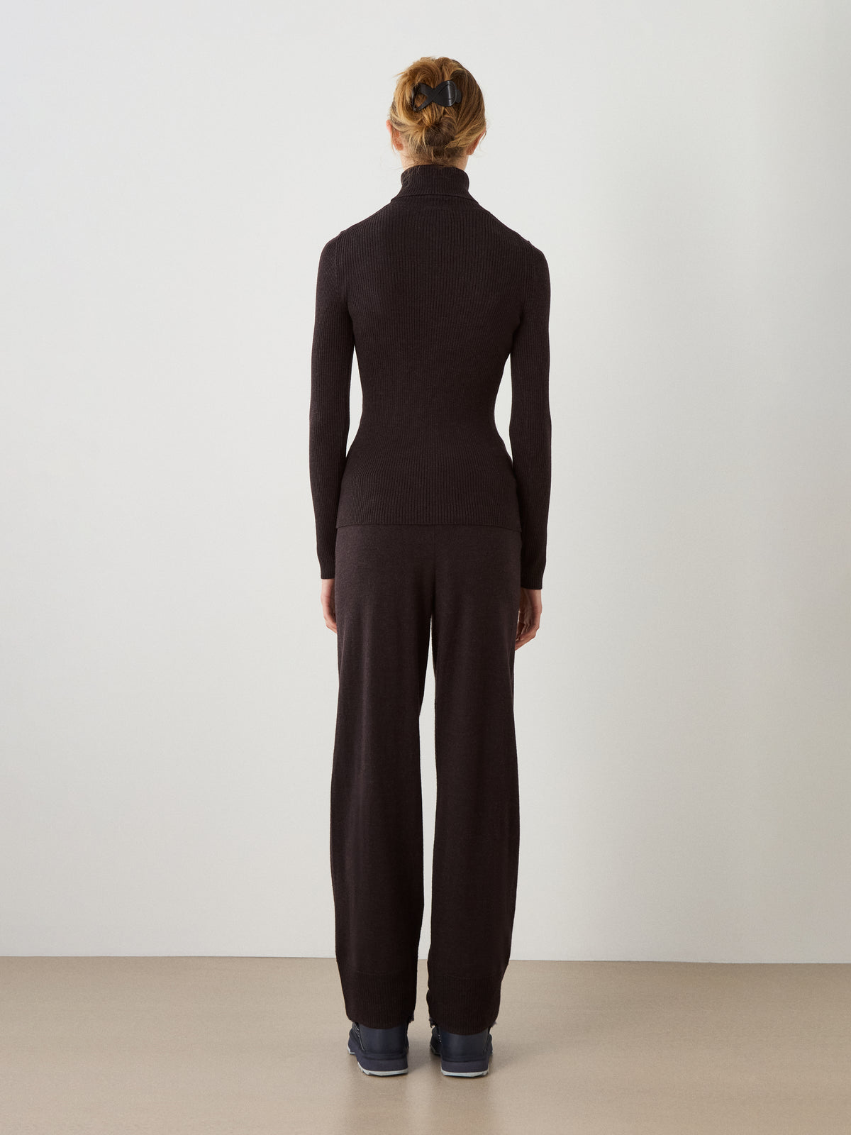 A person with blond hair in a ponytail, wearing the We Norwegians Geilo TurtleNeck for women and black wide-leg pants, stands facing away from the camera against a plain light-colored wall.
