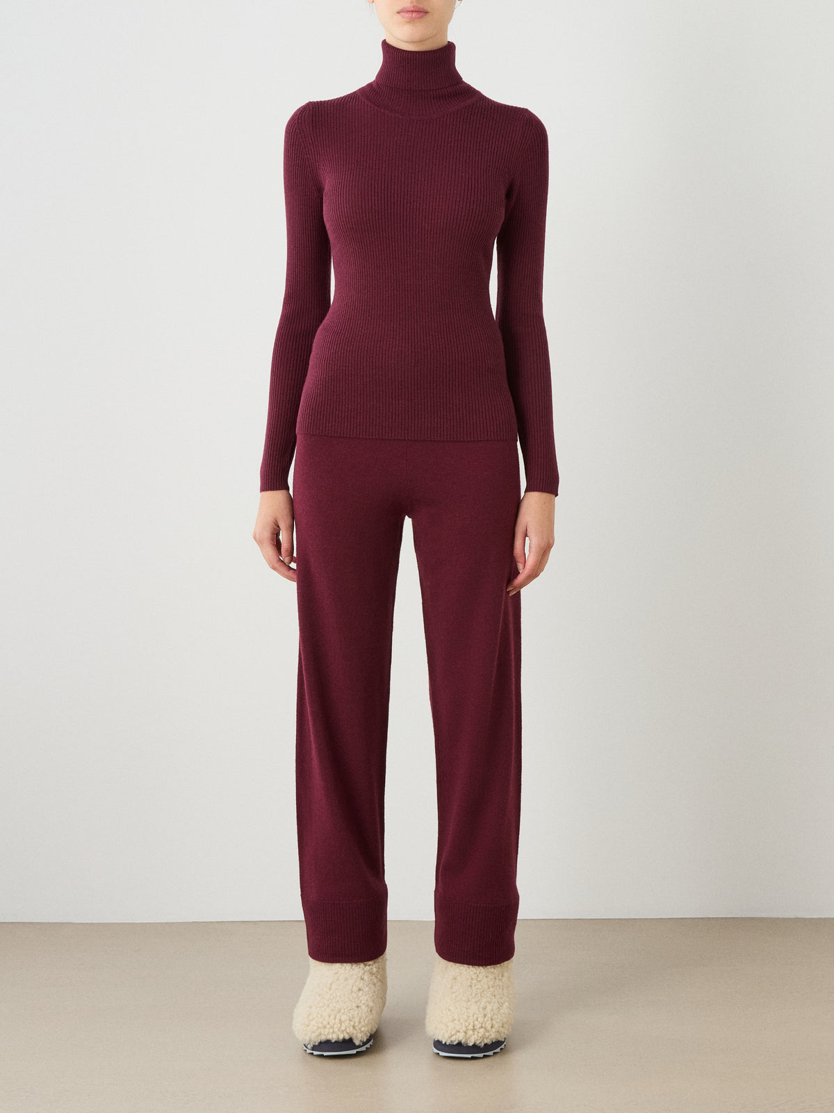 A person is wearing the We Norwegians Geilo TurtleNeck for women in fitted burgundy, paired with matching burgundy pants and cream-colored fuzzy slippers. They are standing against a plain light background, facing forward with their hands by their sides.