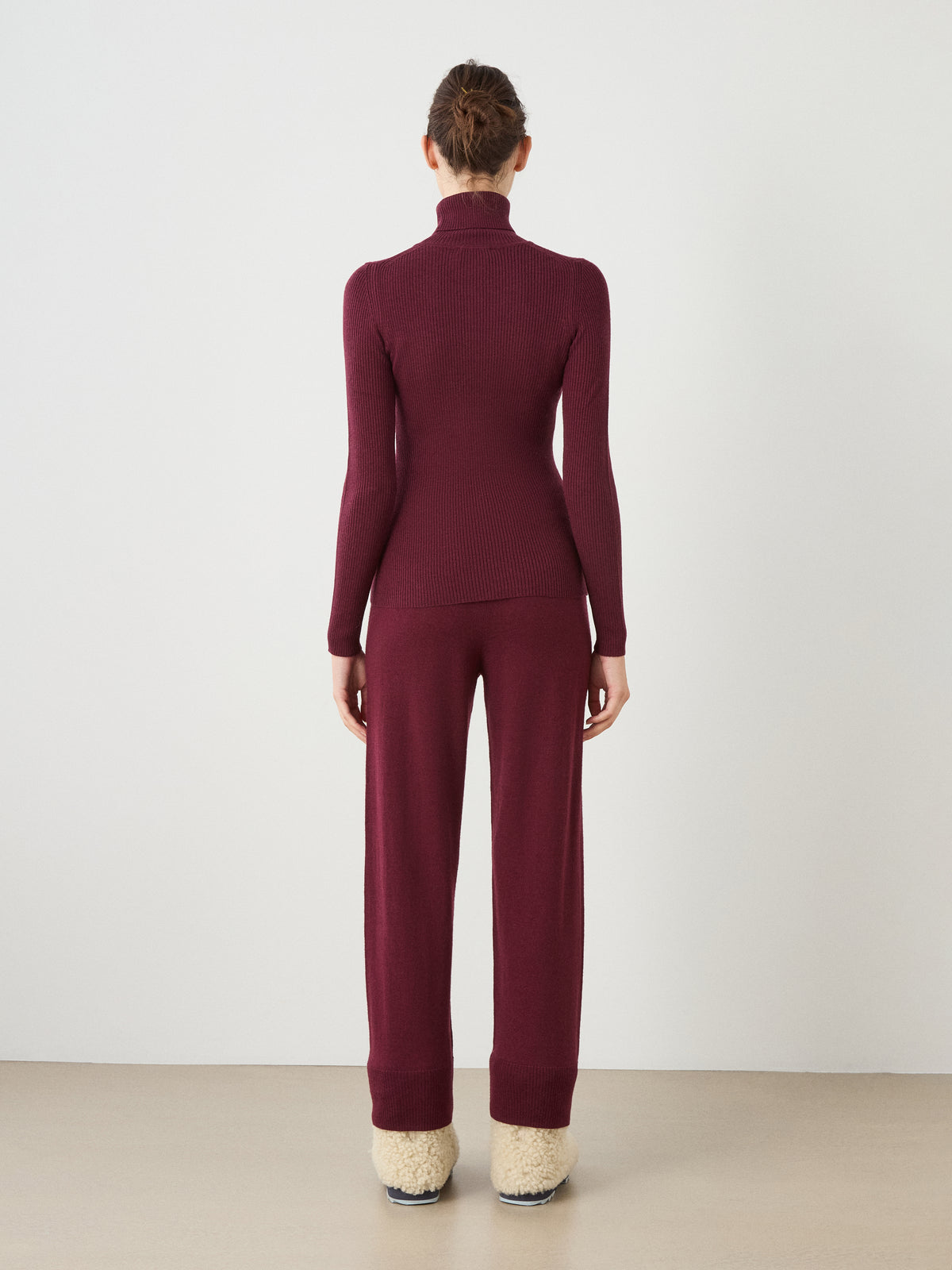 A person stands facing away, wearing the We Norwegians Geilo TurtleNeck for women in maroon, paired with matching maroon pants and fuzzy cream slippers, set against a plain light background.