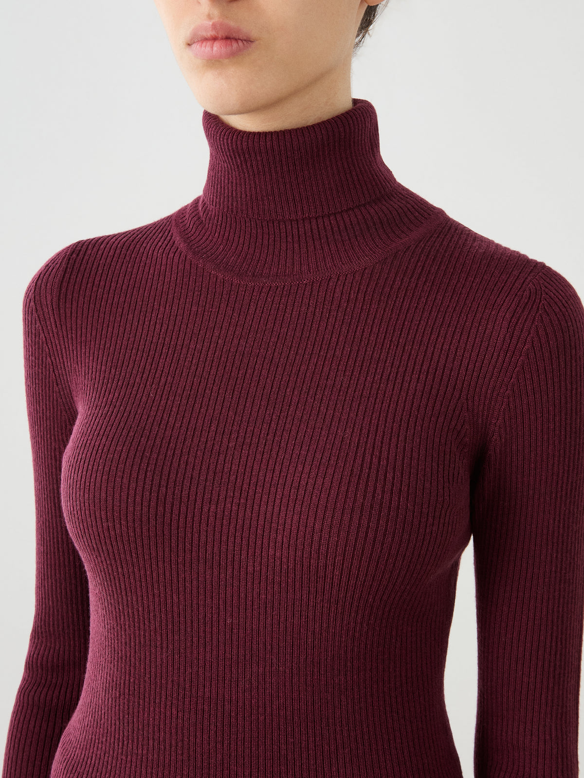A person wearing the We Norwegians Geilo TurtleNeck women sweater in ribbed, burgundy merino wool. The image features the upper torso, neck, and part of the face against a plain light background.