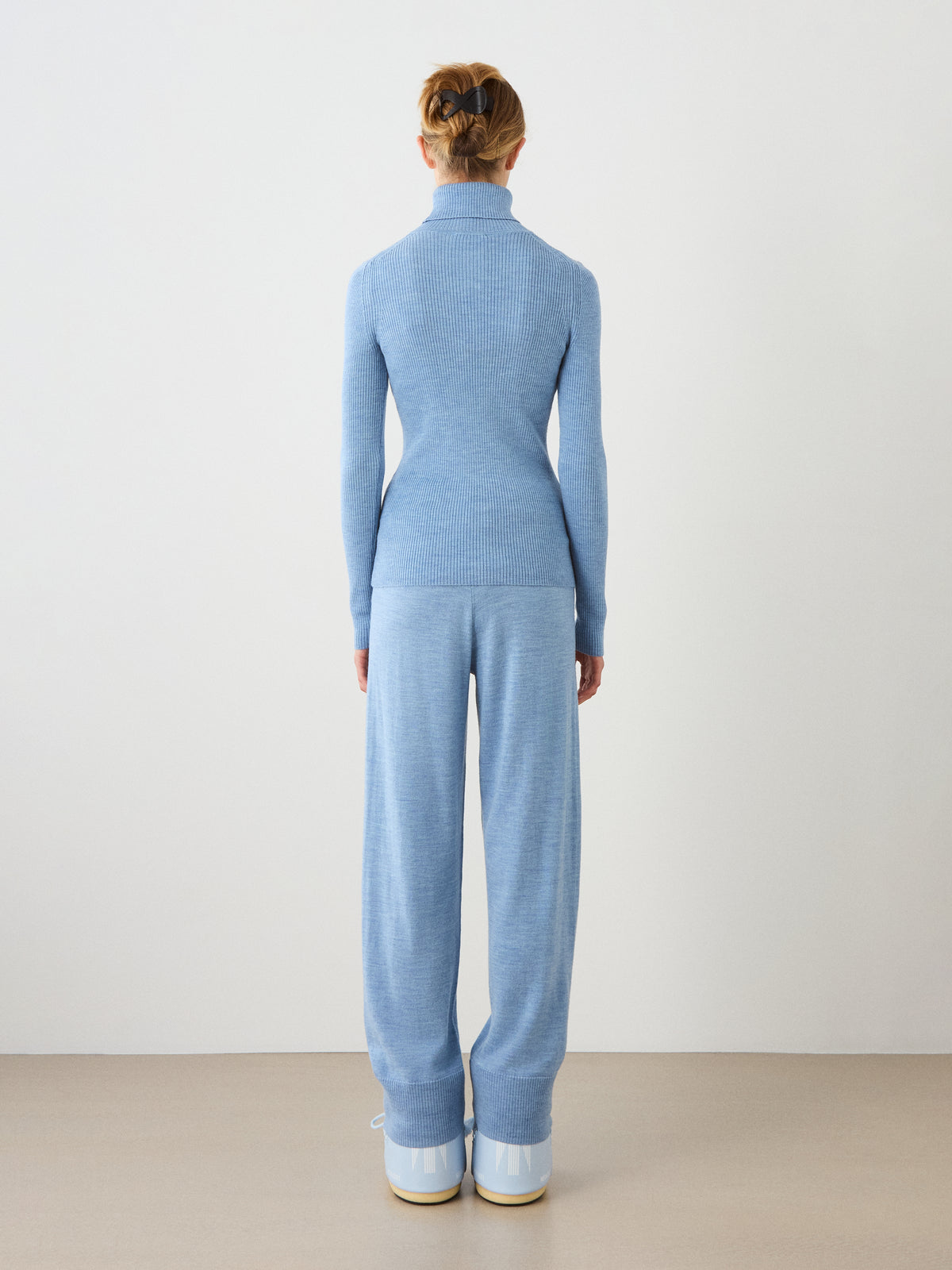 A person stands facing away, wearing the light blue Geilo TurtleNeck women sweater by We Norwegians, with matching loose pants and white platform shoes, against a plain light background.