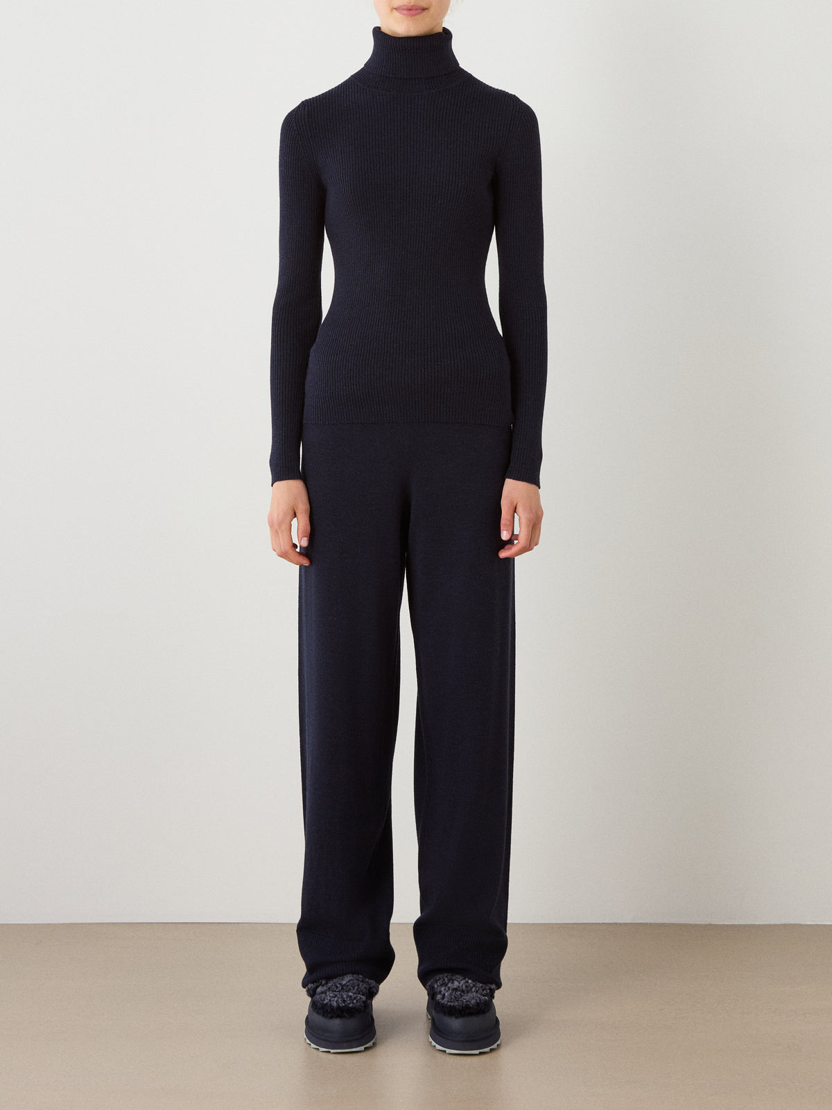 A person stands against a plain wall wearing the We Norwegians Geilo TurtleNeck women’s sweater, paired with loose black pants and dark shoes. Their hands are at their sides, and their face is not visible.