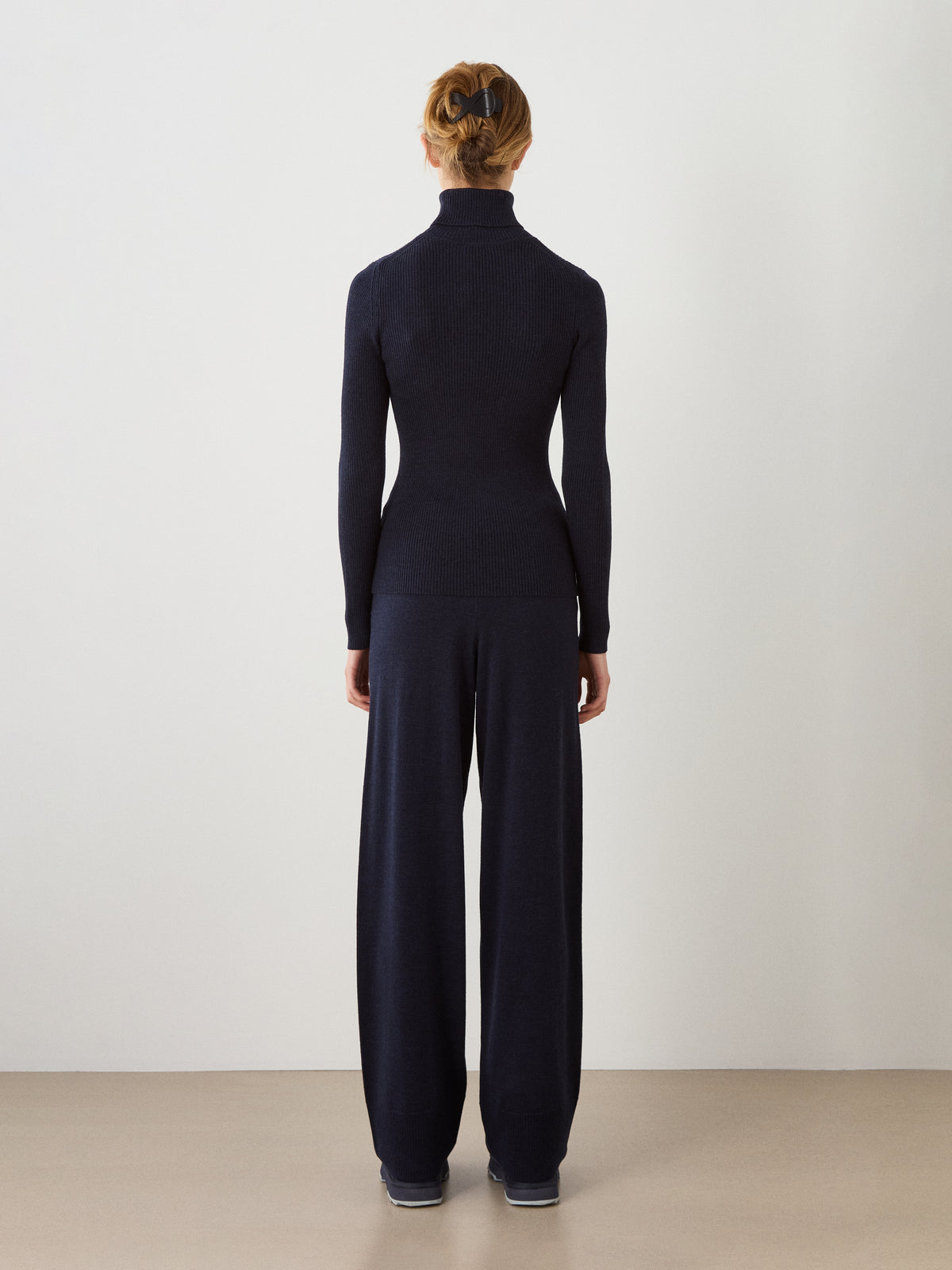 A person stands facing away from the camera, wearing the We Norwegians Geilo TurtleNeck for women in navy, paired with wide-leg pants. Their hair is tied back and their hands are relaxed by their sides, set against a plain white wall and a light-colored floor.