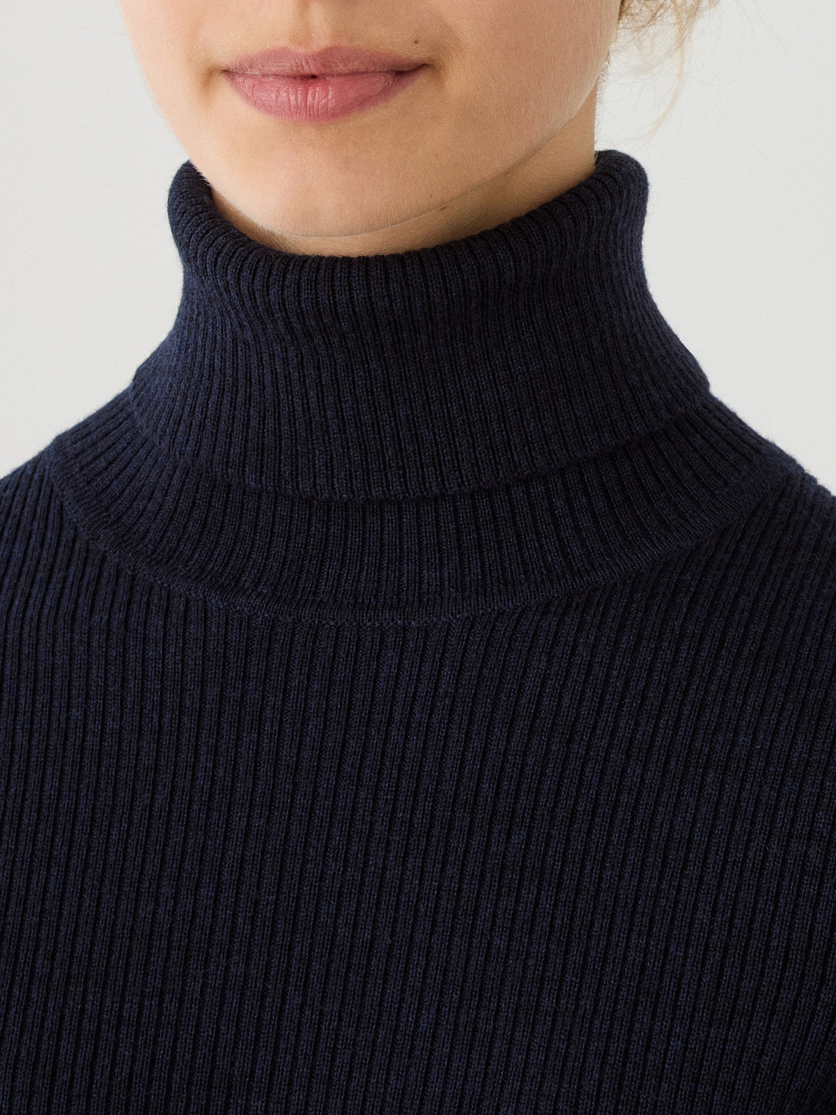 A close-up of a person wearing the We Norwegians Geilo TurtleNeck for women in navy blue merino wool. Only the lower half of the face and neck are visible, highlighting the cozy roll neck and fine ribbed texture of this stylish sweater.