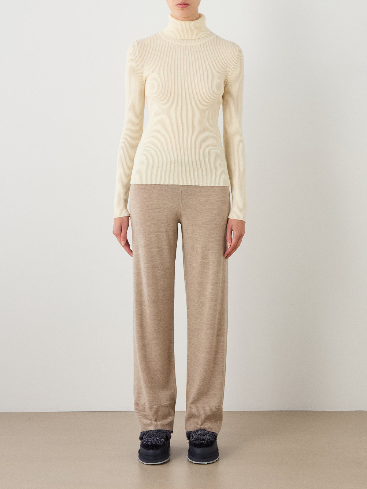 A person stands against a plain wall, wearing the We Norwegians Geilo Turtleneck for women, loose beige pants, and black shoes. Their arms are at their sides and their face is not visible.