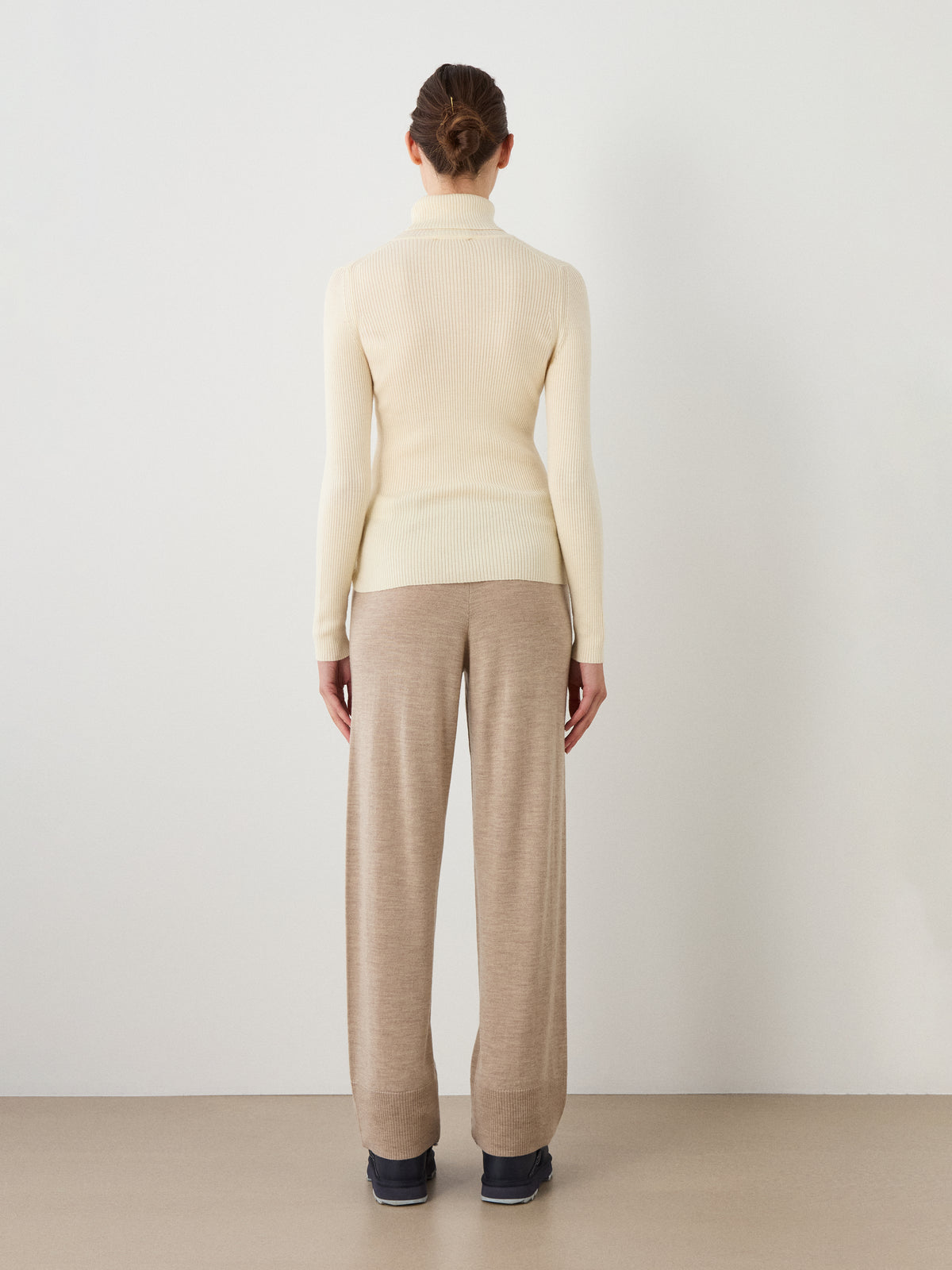 A person stands facing a white wall, seen from behind. They wear the We Norwegians Geilo TurtleNeck women sweater in cream, loose beige pants, and dark shoes. Their hair is tied back in a neat bun.