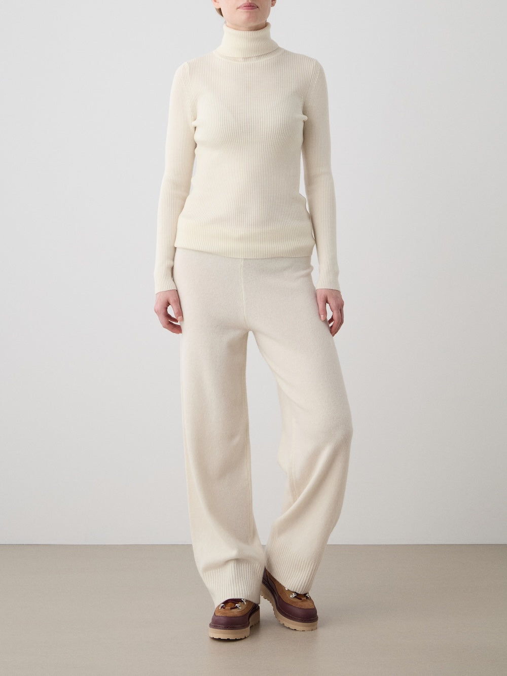 A person stands against a plain background, wearing the cream-colored We Norwegians Geilo TurtleNeck for women with matching wide-leg pants and brown shoes. Their face is partially out of frame.