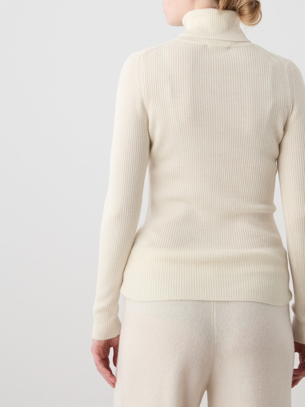 A person stands facing away, wearing the We Norwegians Geilo TurtleNeck women’s sweater in cream with matching light pants against a plain background.
