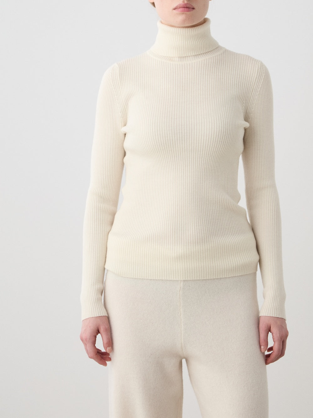 A person models the We Norwegians Geilo TurtleNeck women—a cream merino wool ribbed top—paired with matching light pants against a plain background, with only part of their head visible.