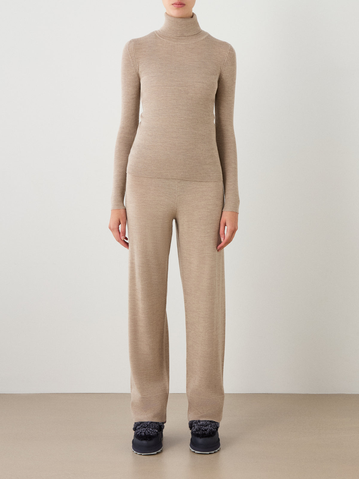 A person stands against a plain wall wearing the We Norwegians Geilo TurtleNeck women sweater in beige, paired with matching beige wide-leg pants and black shoes. Their arms are relaxed by their sides.
