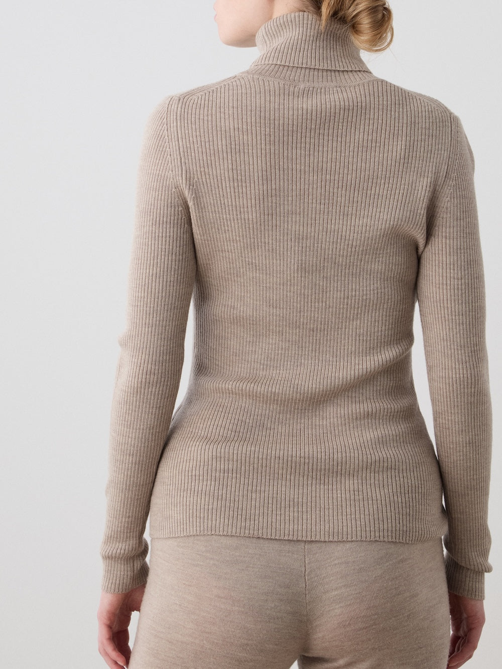 A woman is seen from the back wearing the We Norwegians Geilo TurtleNeck top and matching fitted pants in beige merino wool, standing against a plain light background.