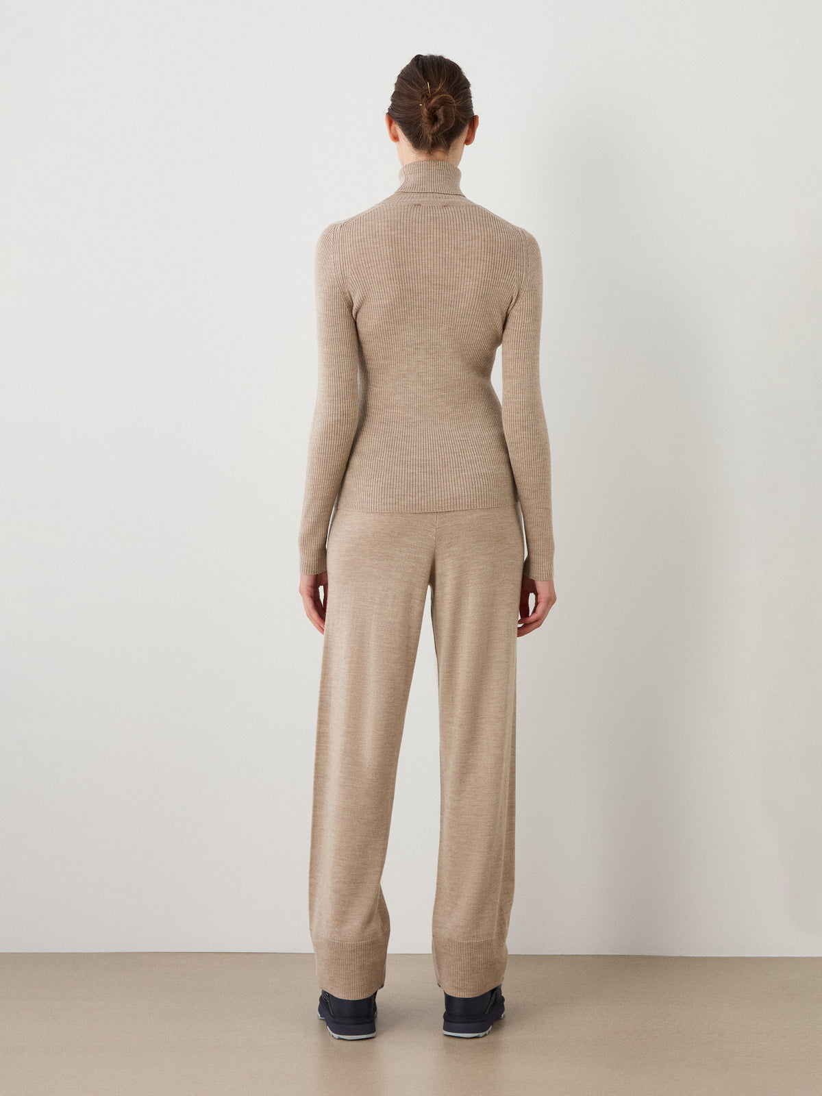 A person with brown hair in a bun is seen from behind, standing against a white wall, wearing the We Norwegians Geilo TurtleNeck women sweater in beige along with matching beige wide-leg pants.