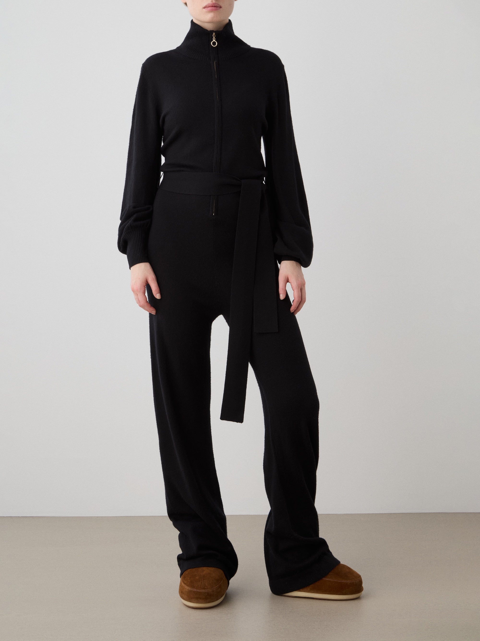 A woman stands against a plain background wearing the We Norwegians Geilo Jumpsuit women in black, featuring long sleeves and a waist tie. Paired with brown slippers, her hands rest by her sides. Her face is not visible. Ideal for a mountain resort look.