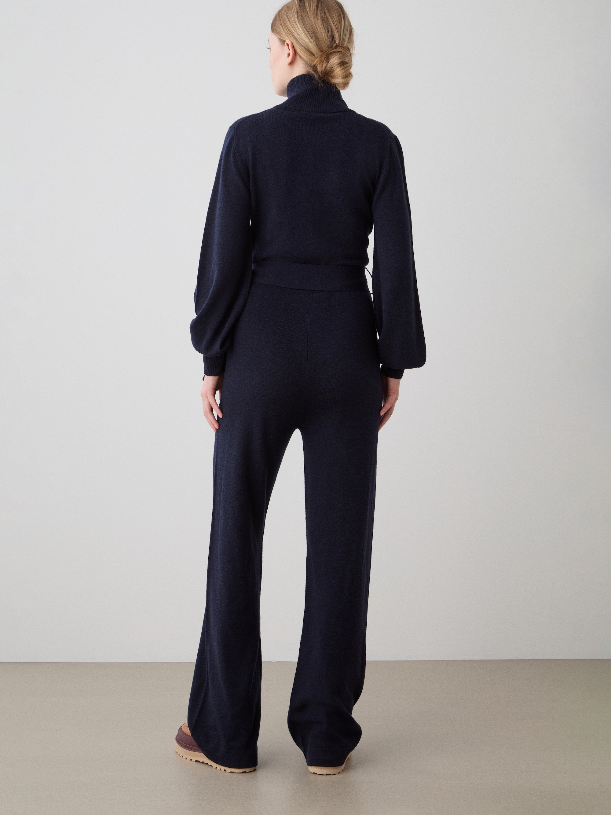 A woman with her hair in a low bun faces away, wearing the We Norwegians Geilo Jumpsuit women in navy—wide legs, long sleeves. She stands on a neutral floor against a plain light wall, showcasing an ideal mountain resort look.