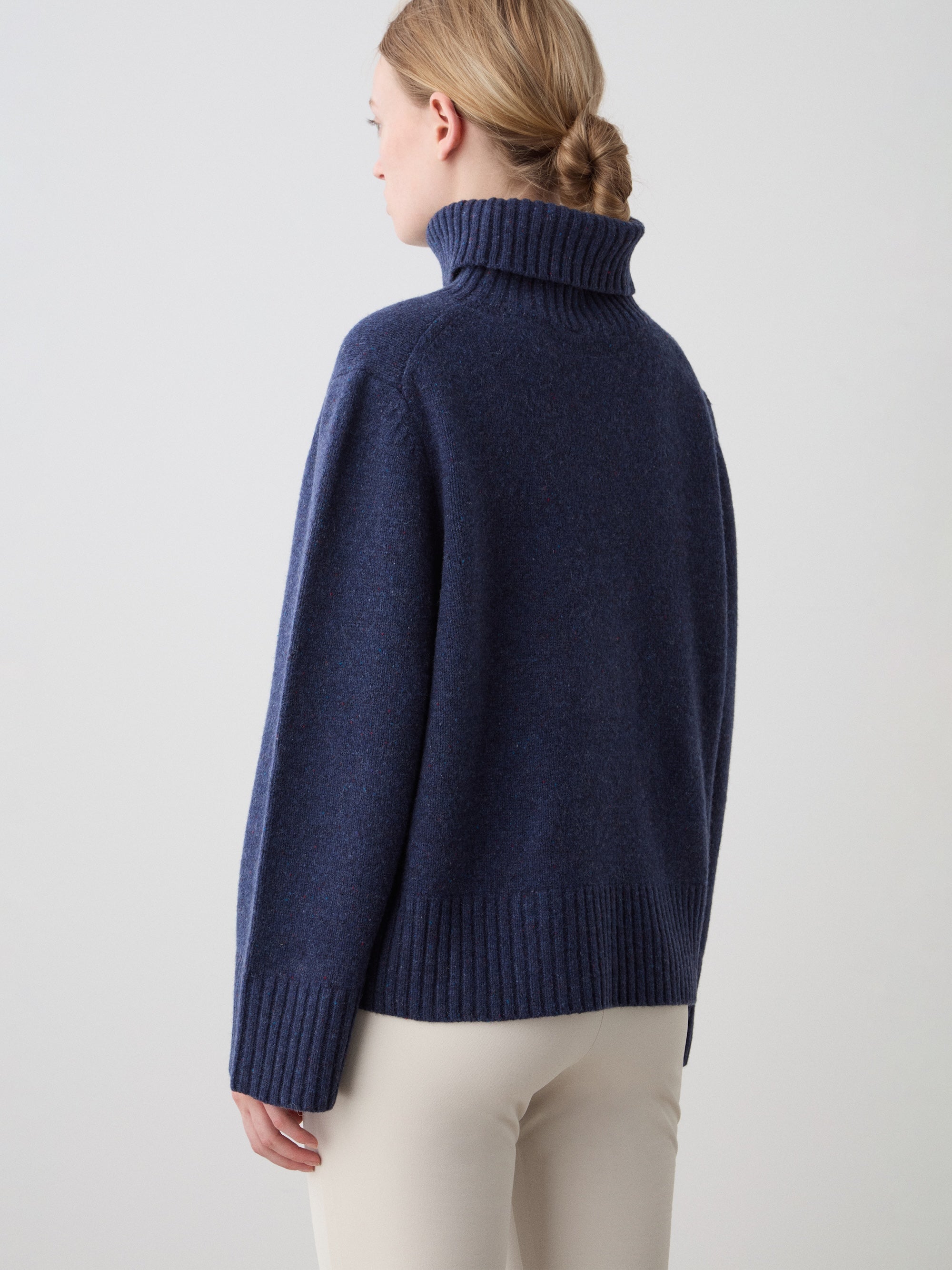 A woman with blonde hair in a low bun stands with her back to the camera, wearing the We Norwegians Highland Sweater for women in navy Extra Fine Merino Wool and light pants, set against a plain light background.