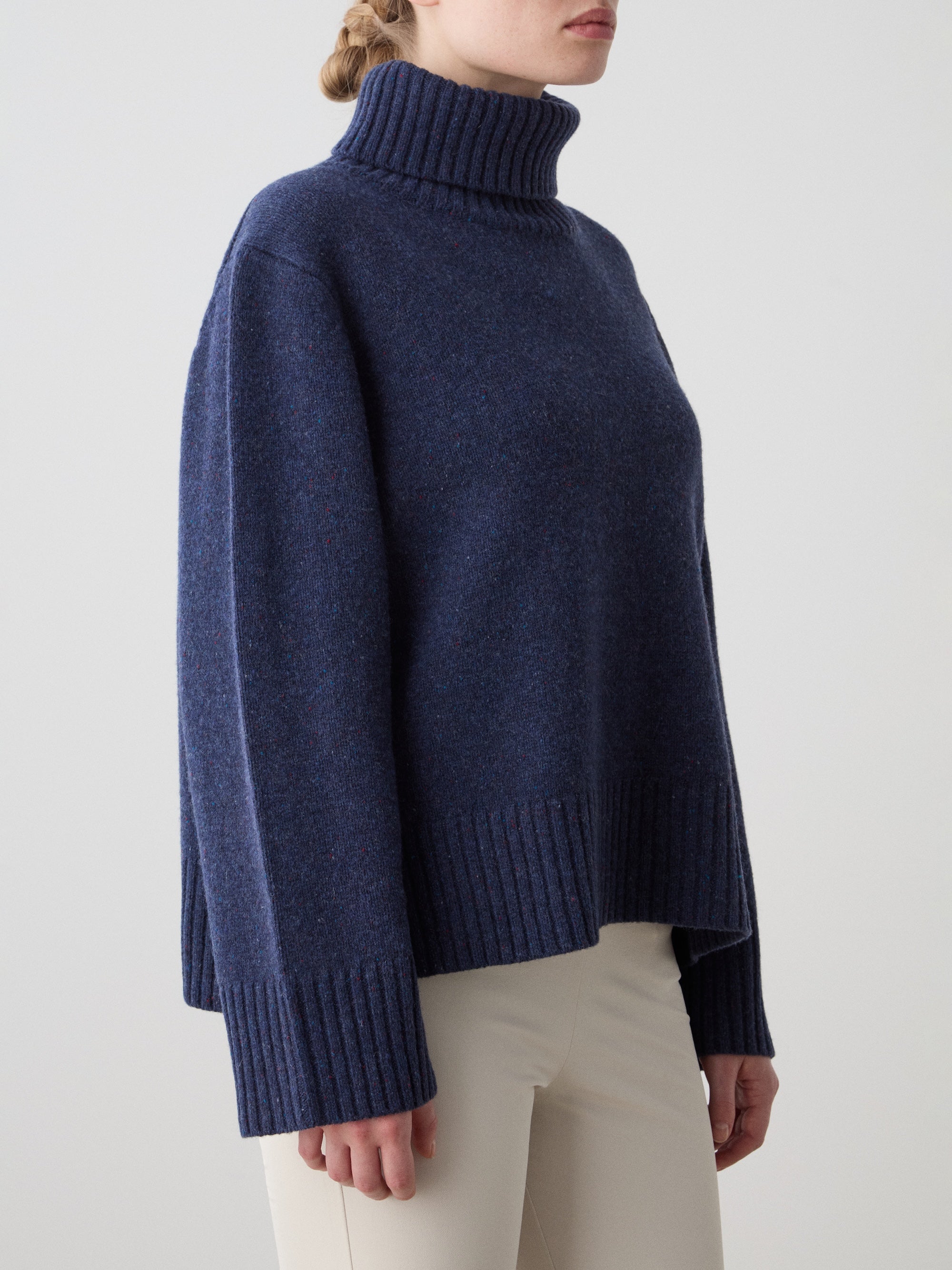 A woman models the We Norwegians Highland Sweater in navy blue—crafted from RWS-certified Extra Fine Merino Wool—with light beige pants, standing against a plain, light background.