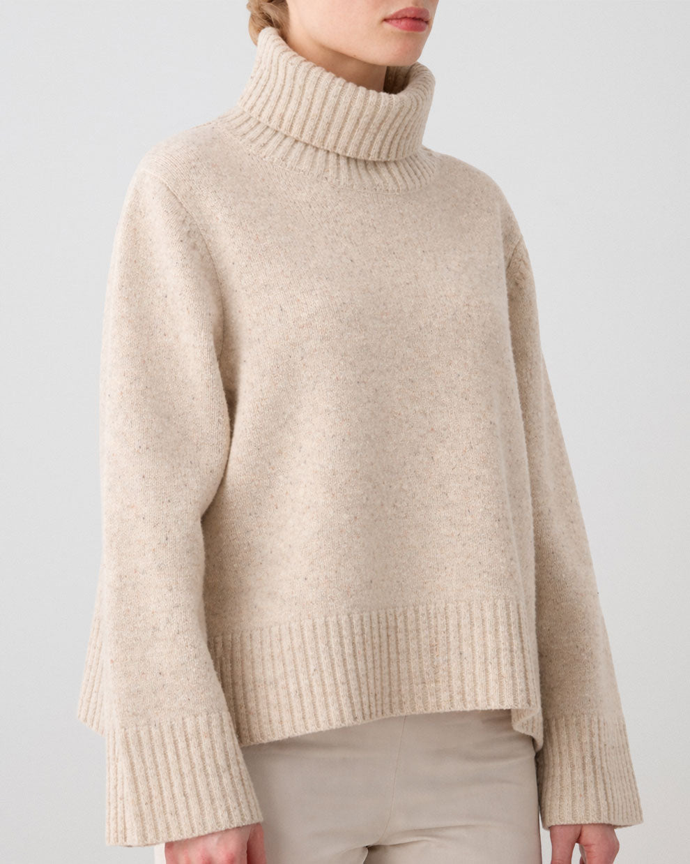 A woman wears the We Norwegians Highland Sweater—a beige, ribbed Merino wool top with wide sleeves—paired with light pants, standing against a plain, light background.