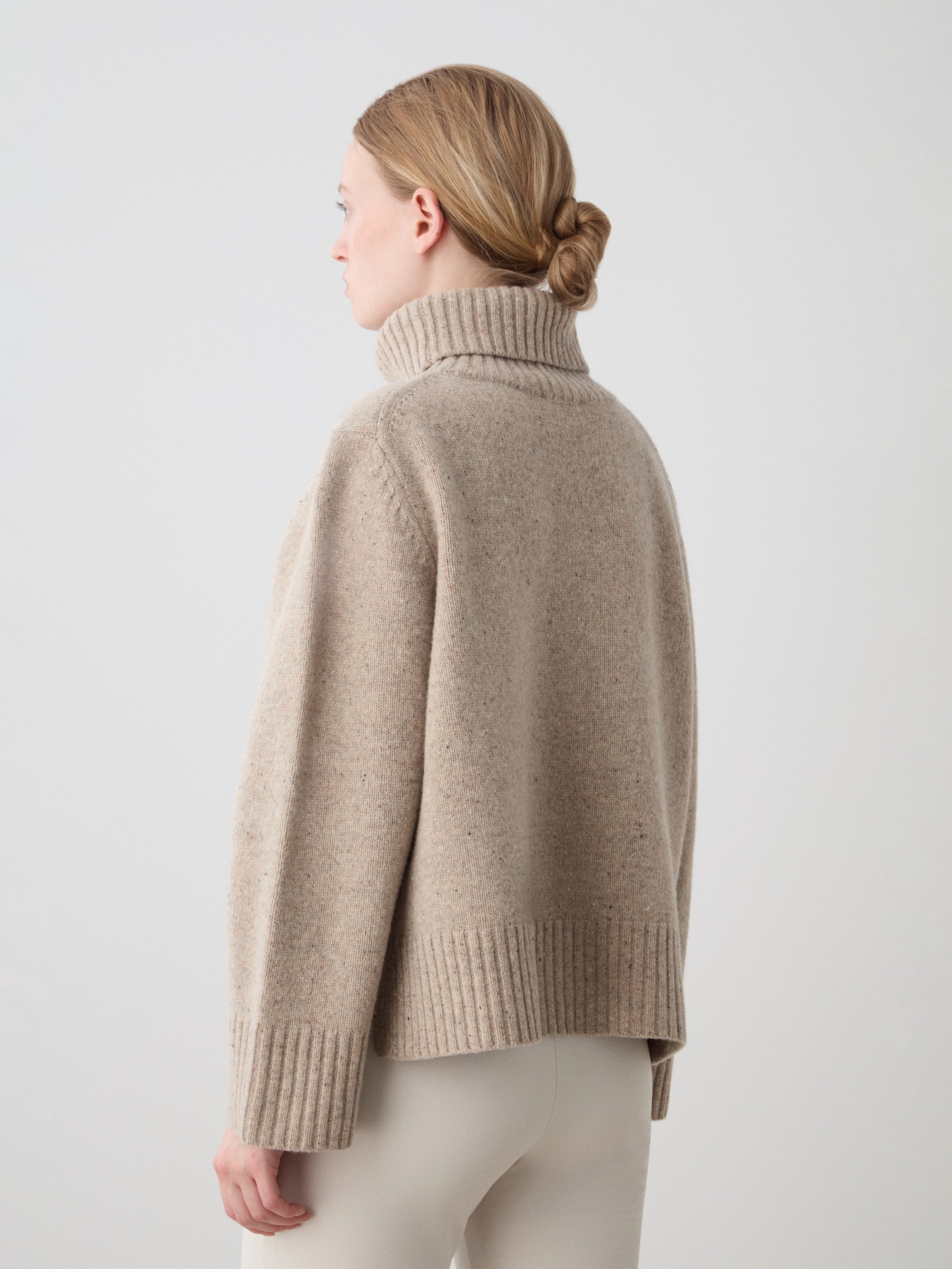 A blonde woman with her hair in a low bun stands sideways, wearing the We Norwegians Highland Sweater for women in beige Extra Fine Merino Wool and light-colored pants against a simple, light background.
