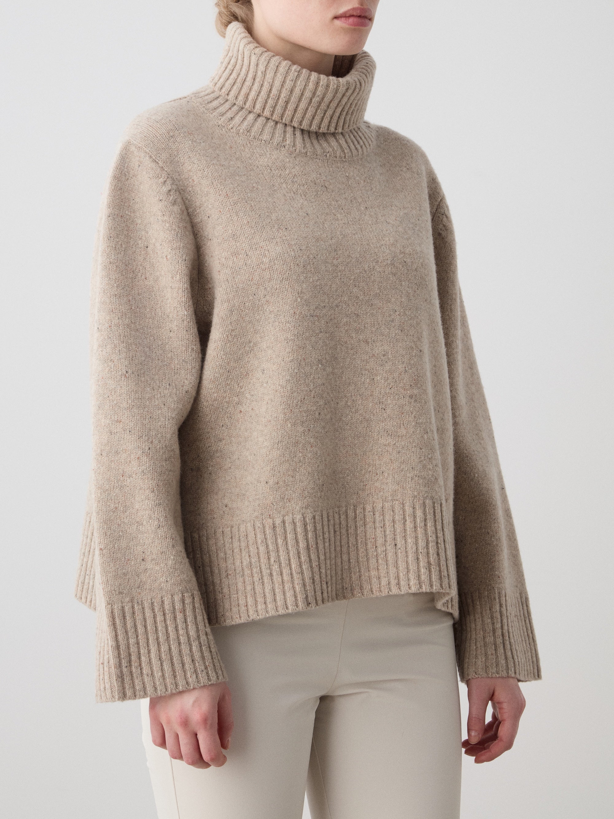 A woman wears the We Norwegians Highland Sweater in beige, made from RWS-certified Extra Fine Merino Wool with ribbed cuffs and hem, paired with light pants against a plain, light background.