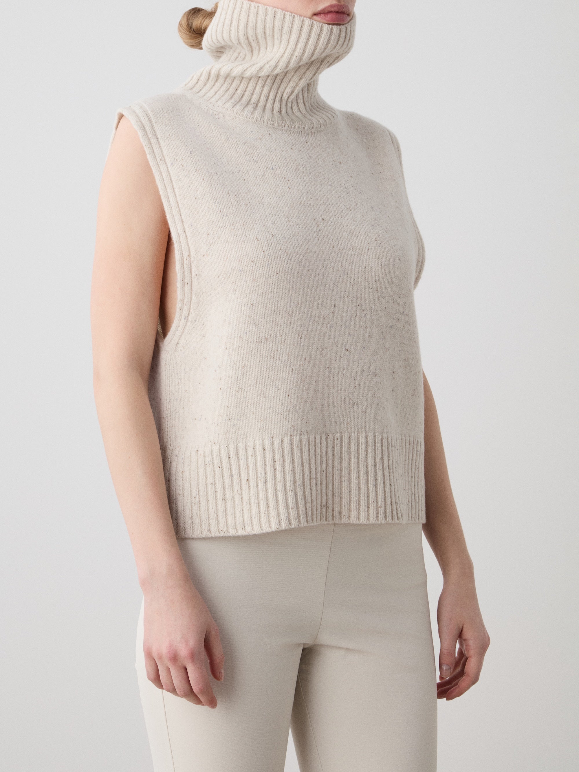 A person wears the We Norwegians Highland Vest for women—a sleeveless, beige merino wool turtleneck—paired with light pants, standing against a plain background. The turtleneck is pulled up to partially cover the mouth.
