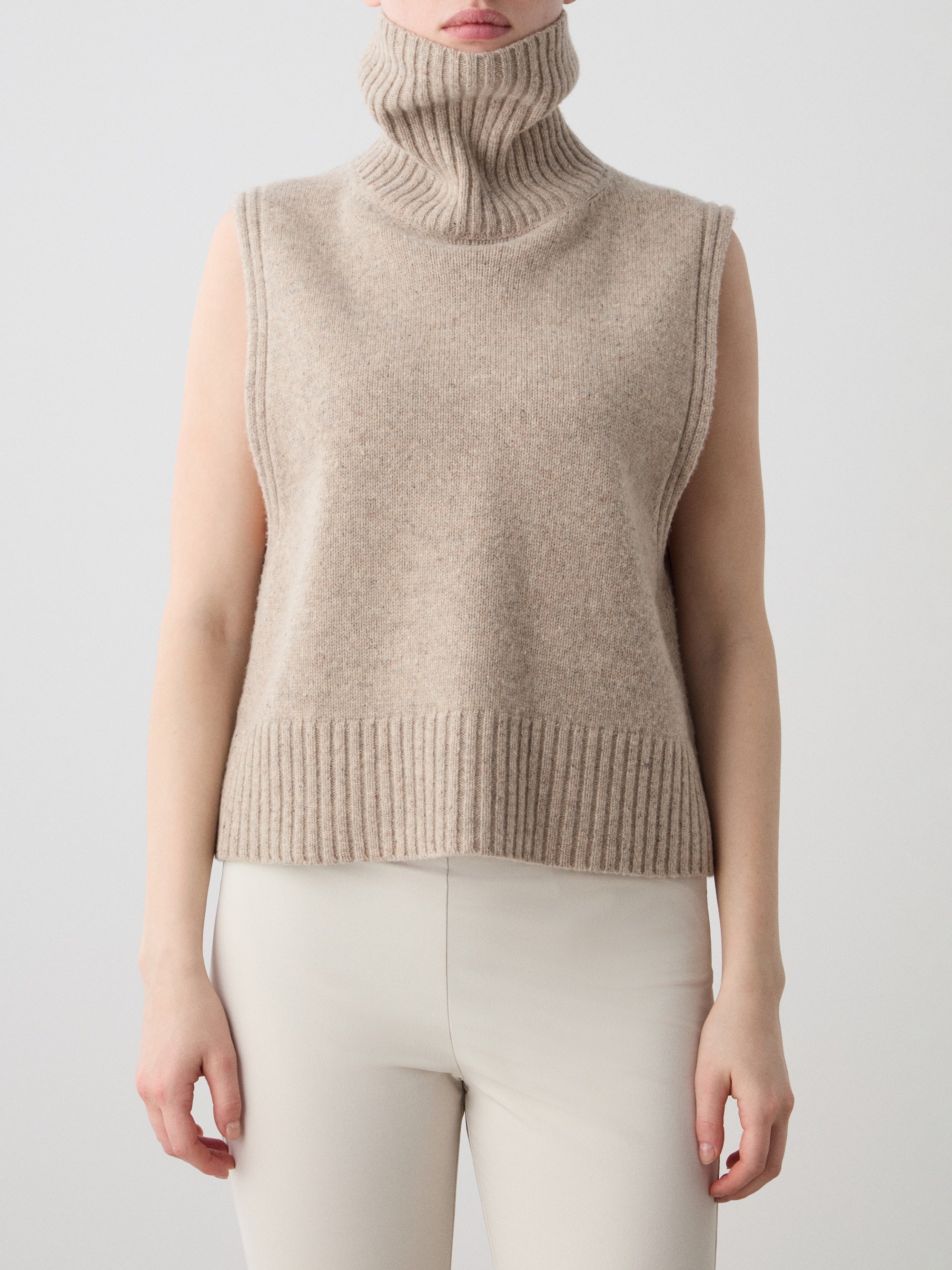 A woman models the We Norwegians Highland Vest, a sleeveless beige merino wool piece with ribbed edges, paired with light pants, standing against a plain light background.