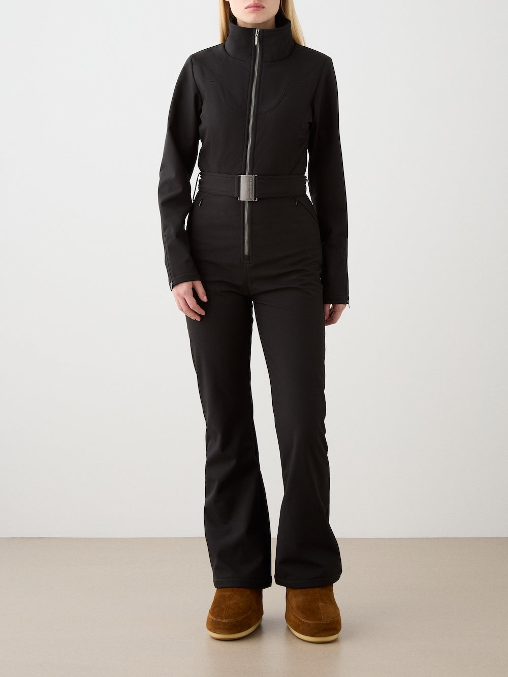 A person stands against a plain background wearing the We Norwegians Western Ski Suit for women—a black zip-up style with a wide belt and brown suede boots. Their hands rest by their sides, and their face is not visible.