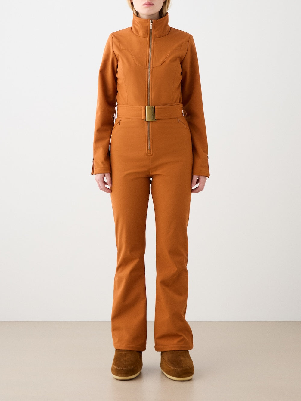 A woman stands facing forward in the We Norwegians Western Ski Suit—fitted, rust-colored with a high collar, belt, and front zipper—paired with tan boots against a plain, light background.