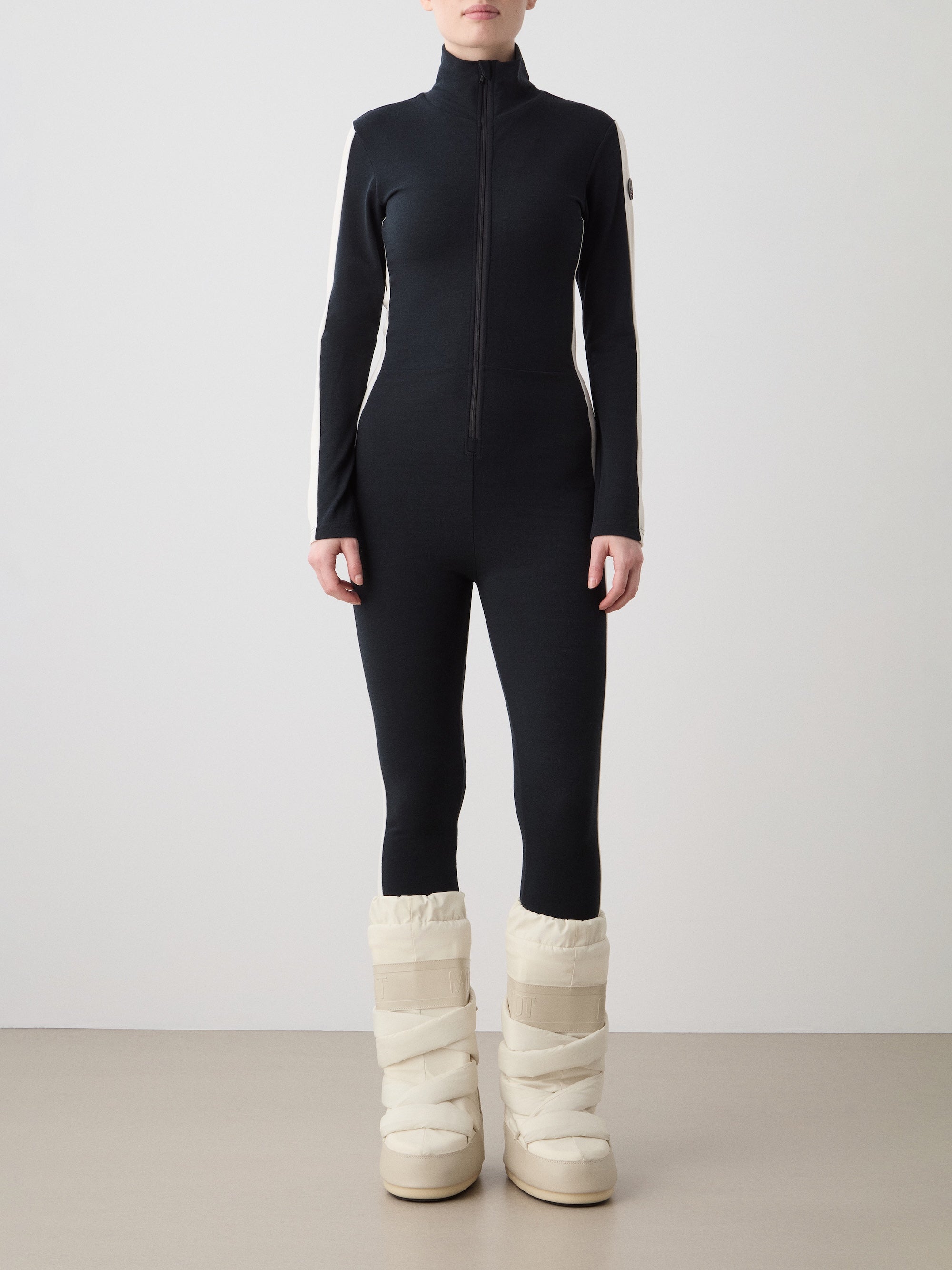 A person faces forward in a fitted black and white Voss Jumpsuit women by We Norwegians, featuring a front zipper, paired with tall padded white snow boots. Their face is not visible against a plain, light background.