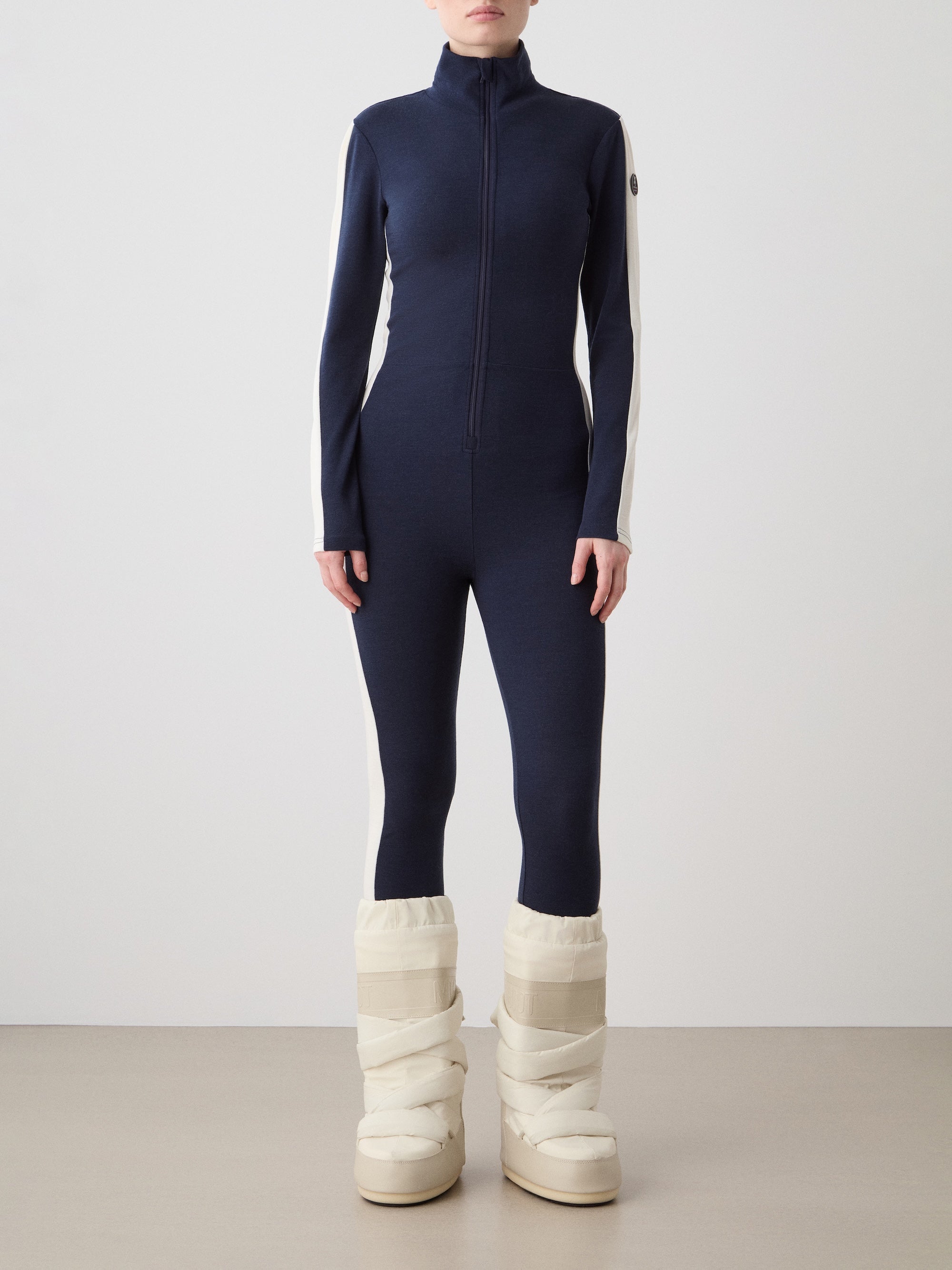 A person stands against a plain background wearing the We Norwegians Voss Jumpsuit for women in navy blue and white, featuring a front zipper and high collar, paired with cream quilted snow boots. The face is not visible.