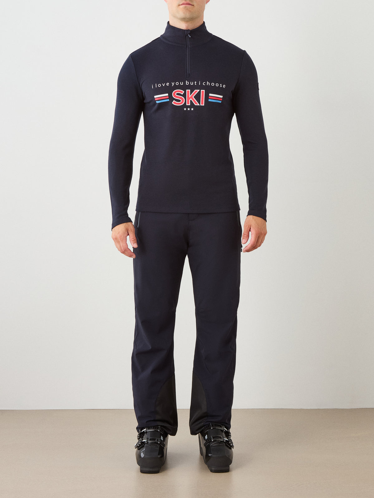 A person stands facing forward wearing the We Norwegians Moore ZipUp men ski top—black, vintage-inspired, with “I love you but I choose SKI” across the front—paired with black pants and black ski boots, set against a minimalist white and beige backdrop.