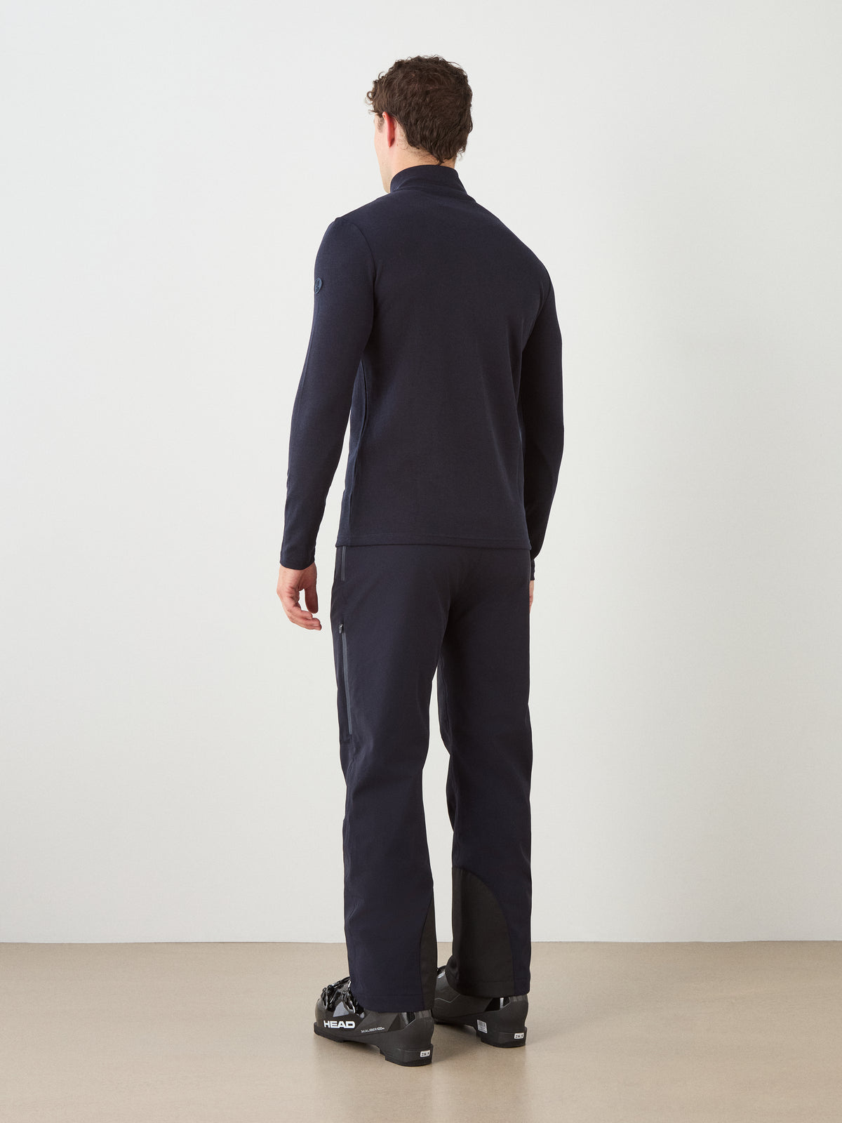 A person stands facing away from the camera, wearing the We Norwegians Moore ZipUp men set and black ski boots, in a minimalist indoor setting with a light-colored floor and plain white wall.