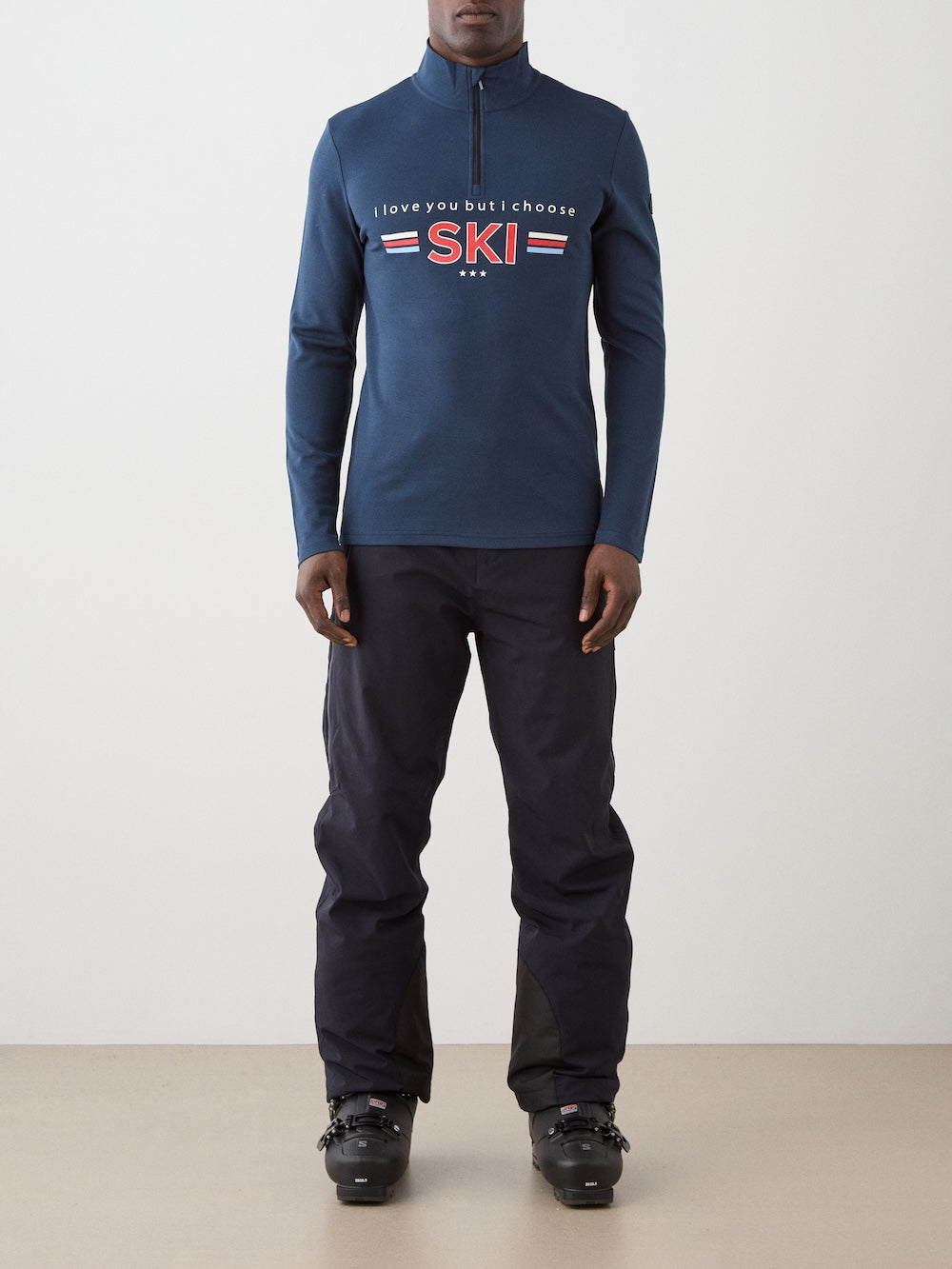 A person wearing the We Norwegians Moore ZipUp men blue retro ski base layer with "I love you but I choose SKI" print, black pants, and black ski boots, standing indoors against a plain light background.